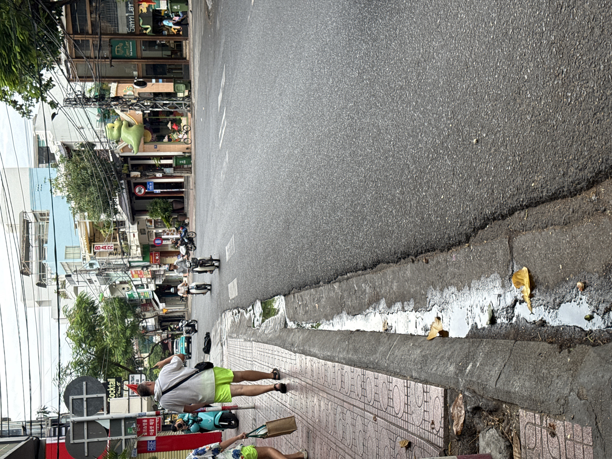 A street scene in Nha Trang, Vietnam, depicts a grey asphalt road with a concrete drainage ditch running along the side. Several people are visible, including a man in a white t-shirt and bright green shorts walking on the sidewalk, facing away from the camera. He is wearing a red party hat and has a black bag slung over his shoulder. Another person, partially visible on the left, is walking on the sidewalk with a shopping bag. Several motorbikes are present on the road, with some riders in motion. Buildings line the street, featuring various signage and architectural styles, including a building with a prominent green cartoon character decoration. Overhead, a complex network of electrical wires crisscrosses the sky. The ground on the sidewalk has a patterned tile surface, and the drainage ditch contains water and fallen leaves.