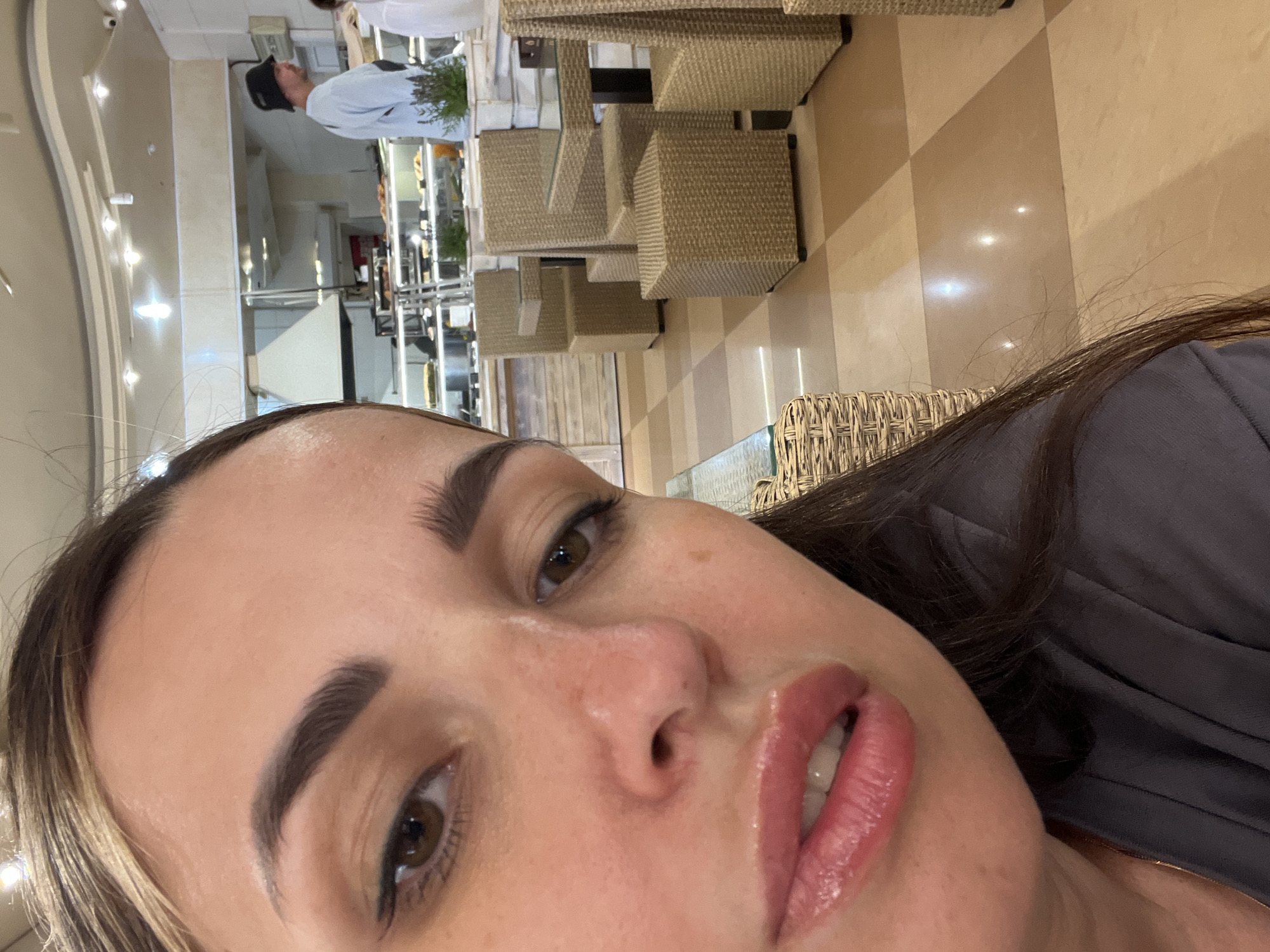 The image is a selfie taken by a woman in what appears to be a cafe or restaurant. She is close to the camera, with her face taking up most of the frame. Her hair is dark brown and pulled back, with some strands falling loose around her face. She is wearing makeup, including eyeliner and lip gloss. Her lips are slightly parted, revealing her teeth.
In the background, there are tables and chairs made of woven material, suggesting an outdoor or conservatory setting. There are also displays of food and what looks like a kitchen area, with a man in a black hat visible. The floor is tiled in a checkered pattern of beige and brown. The overall lighting suggests an indoor environment with artificial light sources. The location is described as Unknown, Unknown.