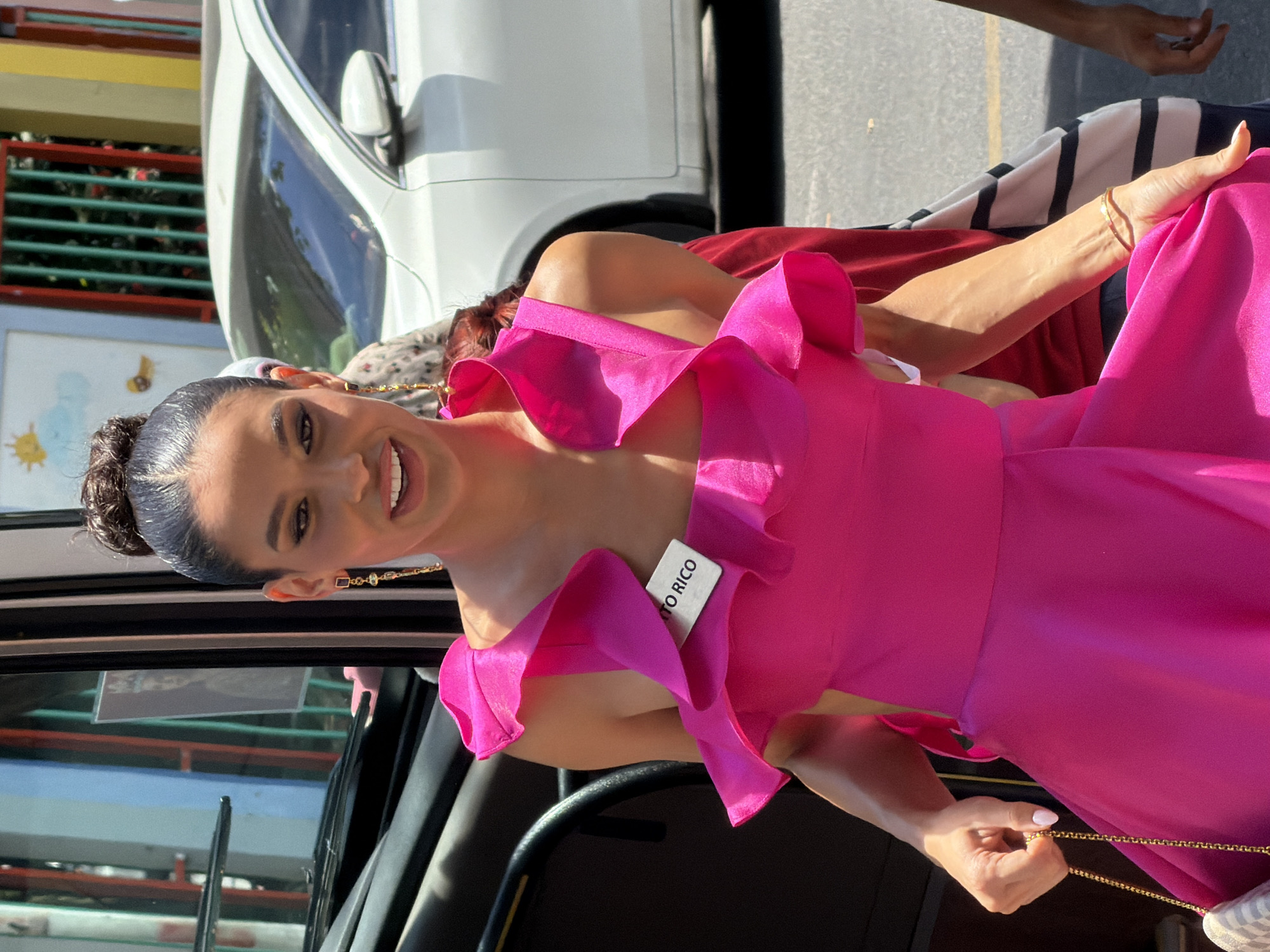 A woman wearing a bright pink, ruffled dress and a "PUERTO RICO" tag on her chest is captured in a medium shot outdoors. She has her dark hair pulled up in a bun and is wearing dangling earrings. Her mouth is slightly open, and she appears to be smiling or speaking. She is holding a small, checkered purse with a gold chain in her left hand. A white car is visible in the background, partially obscuring a fence with vertical green bars. The environment suggests a public outdoor setting, possibly a street or an event area. The image does not contain any specific visual cues that would indicate the location as Nha Trang, Vietnam.