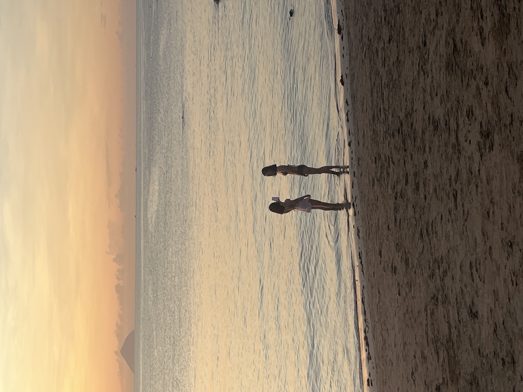 Two figures stand on a beach in Bac Nha Trang Ward, Vietnam, where the shoreline meets the ocean at sunset. One figure holds a phone. The sky is bright, with an orange hue near the horizon. The sandy beach is to the right.