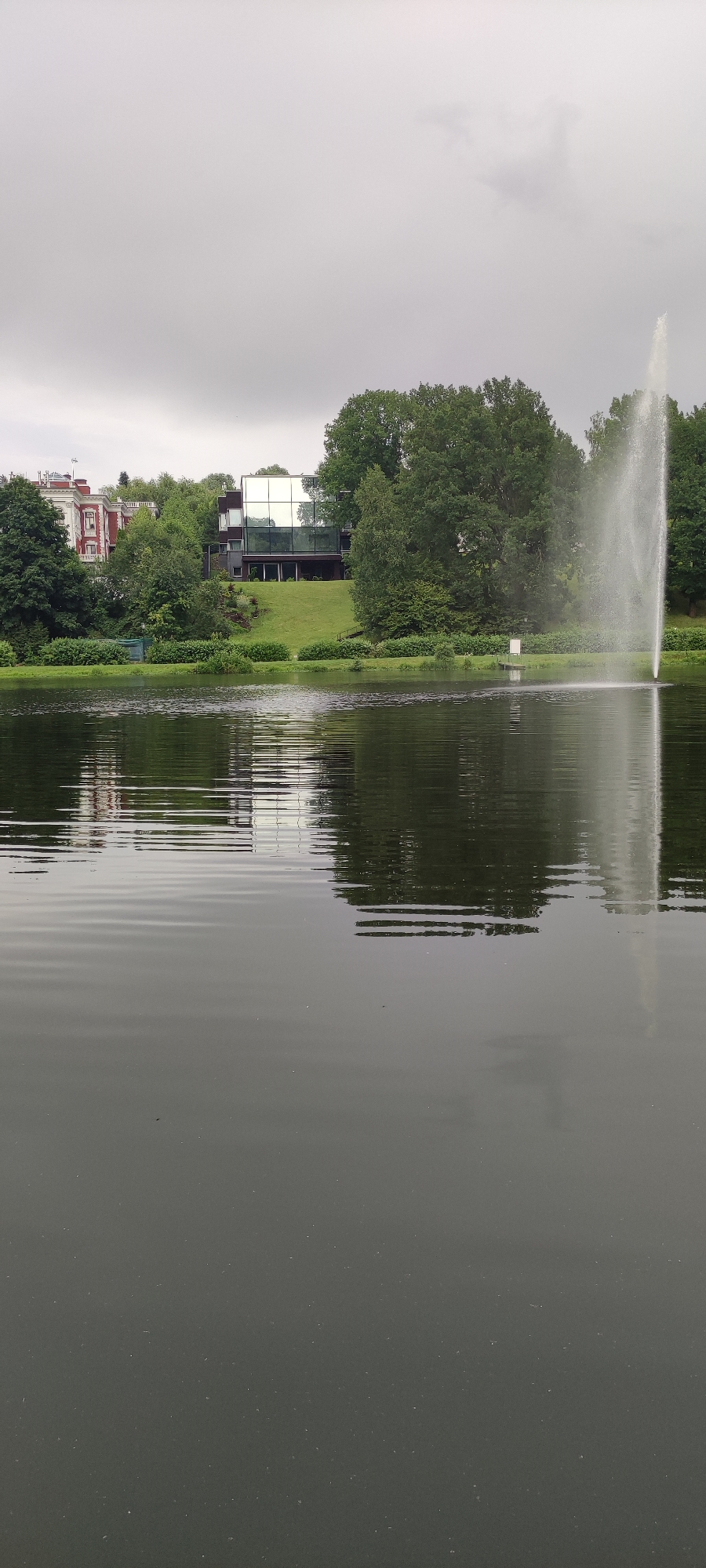 The image depicts a serene lake scene with a modern glass-fronted building and a fountain on the far bank. To the left, a more traditional red-brick building is visible, also nestled amongst lush greenery. The sky is overcast, casting a soft, diffused light over the landscape. The water of the lake is calm, with gentle ripples reflecting the surrounding trees and buildings. The overall impression is one of tranquility and understated elegance. This scene is located in Angelovo, Russia.