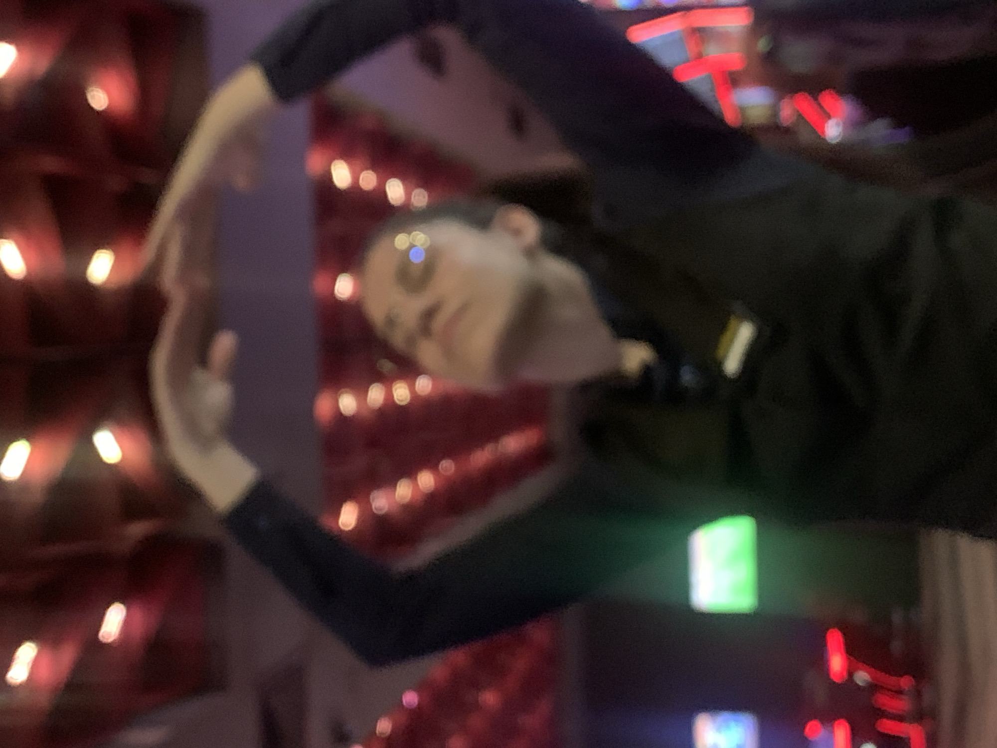 The image depicts a blurred, dimly lit indoor environment, likely a casino or entertainment venue, with a prominent blurry subject in the foreground. The subject, a person wearing dark clothing and a vest with a yellow tag, is positioned centrally and is looking upwards with their arms raised above their head, forming a curve. The background features out-of-focus red and purple lights, possibly from decorative lighting or signage. There are also some blurred green and blue lights visible on the left side of the frame. The overall impression is that of a candid shot taken in a low-light, dynamic setting. The specific location in Burgas, Bulgaria is not ascertainable from the visual information present.