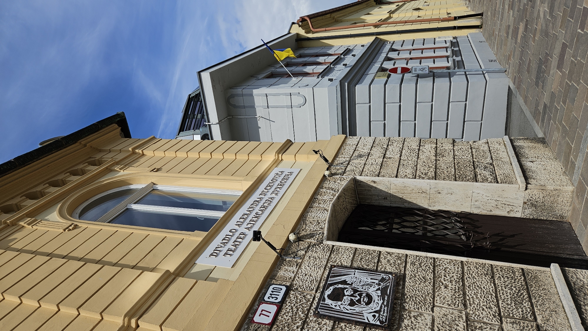 The image shows the exterior of the Alexandr Duchnovič Theatre in Prešov, Slovakia. The theatre is a yellow, two-story building with a light-colored stone facade at the entrance. Above the entrance, a sign reads "DIVADLO ALEXANDRA DUCHNOVIČA" and "ТЕАТЕР АЛЕКСАНДРА ДУХНОВИЧА". To the left of the entrance, there is a portrait of a man and two street signs, one with the number "77" and another with "3110". A dark metal gate is visible over the entrance. To the right of the theatre, a light gray building is visible, with a Ukrainian flag flying from its facade. The street in front of the buildings is paved with bricks. The sky is blue with scattered clouds.