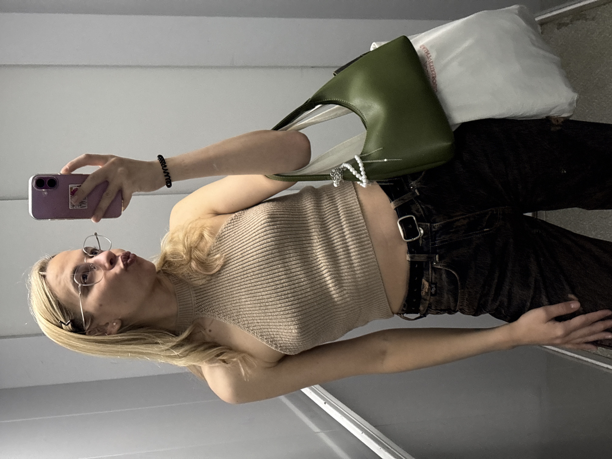 A young woman with blonde hair, wearing glasses, a light brown tank top, and dark pants with a black belt, is taking a selfie. She is holding a purple smartphone in her left hand. Over her shoulder, she carries a green purse and a white shopping bag. The background appears to be an elevator interior with gray paneling. There is no identifiable information to determine if it is located in Krasnoyarsk, Russia.