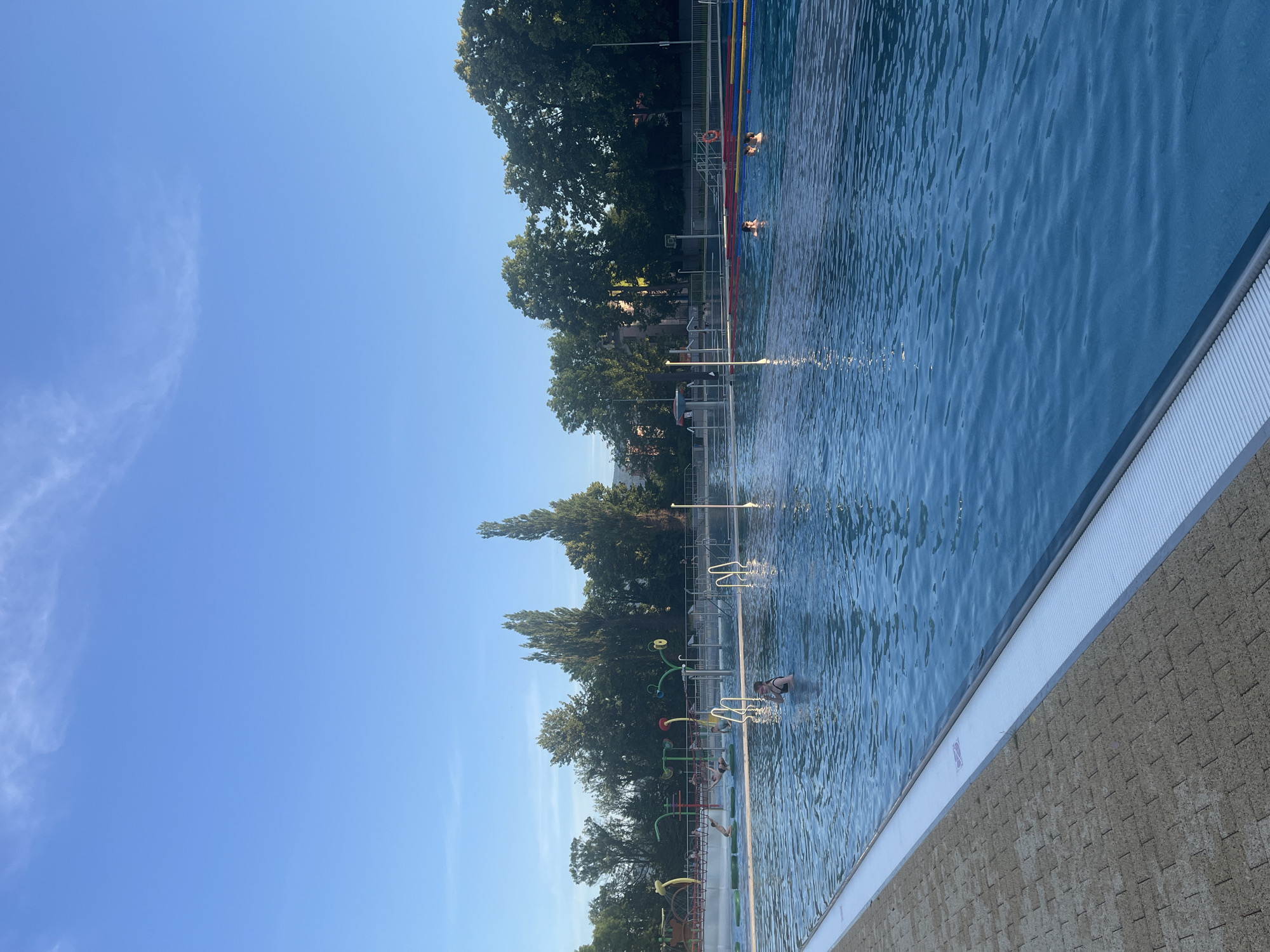 An outdoor swimming pool on a sunny day. The pool has clear blue water and is bordered by a textured tan and grey paved area. Several people are in the pool, some swimming and others standing. In the background, there are trees and buildings under a clear blue sky with wispy clouds. There are also pool features like diving boards and water sprayers.