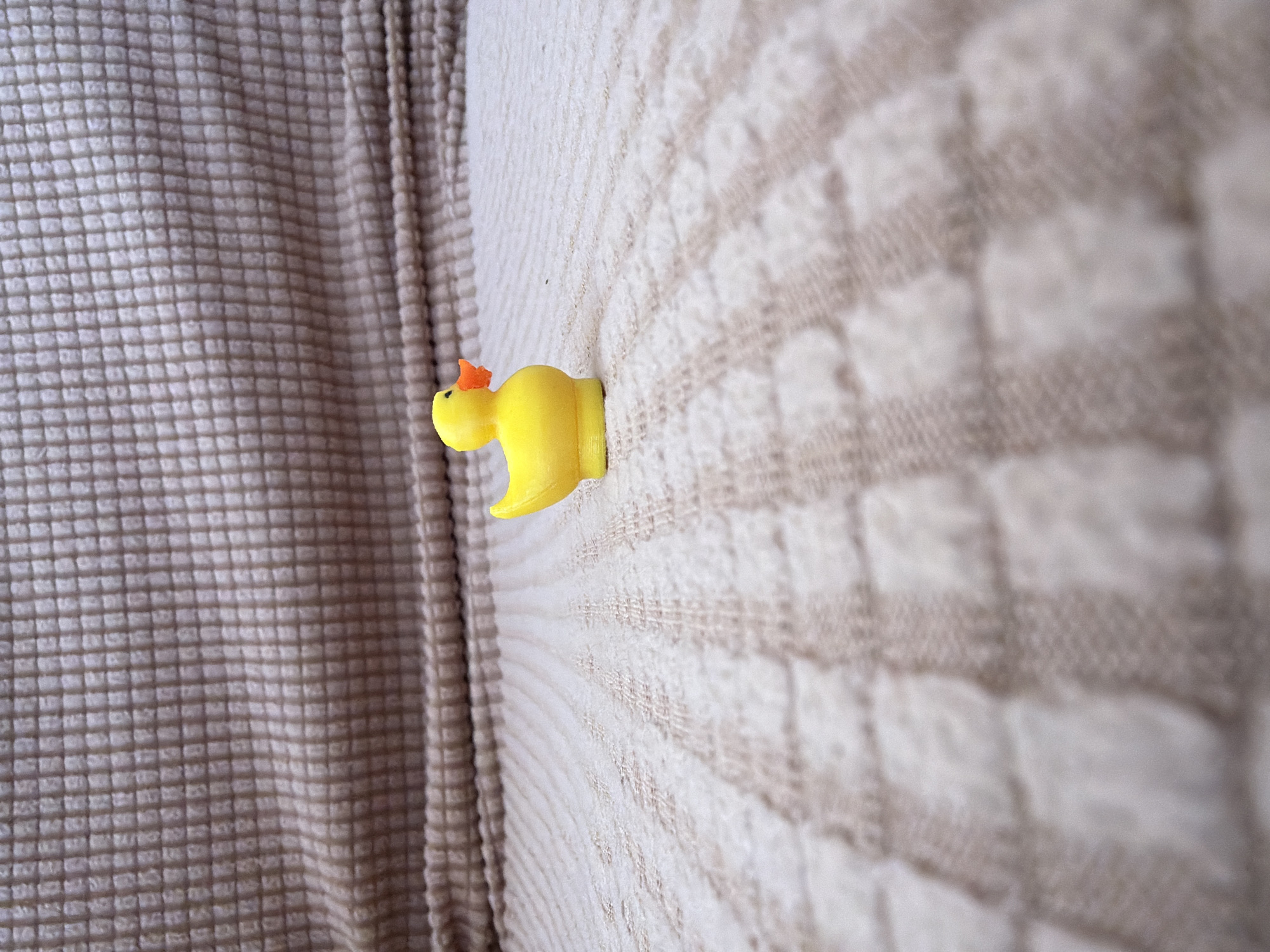 A small, yellow rubber duck toy is positioned in the center of a textured, light brown fabric surface. The duck is facing towards the right side of the frame, with its orange beak slightly angled upwards. The fabric has a repeating checkered pattern, with some areas appearing smoother and others more raised. In the background, a darker, more densely textured fabric is visible, creating a contrasting backdrop. The lighting appears to be soft, with the main focus on the duck and the immediate foreground. The scene is devoid of any people or any indication of a specific location within the city of Япрыково, Russia.