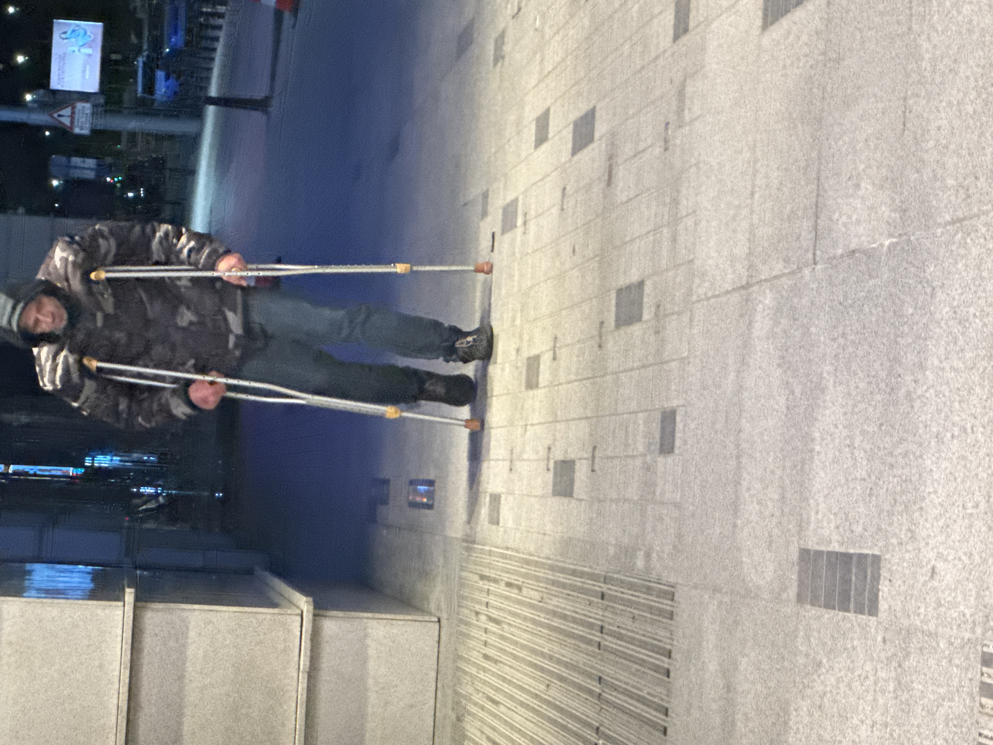 A man using crutches is depicted walking along a sidewalk in Sofia, Bulgaria. He wears a camouflage jacket, dark pants, and shoes, with a gray hat covering his head. The sidewalk is composed of gray stone tiles with some square insets and grooved sections. In the background, city lights are visible, along with a signpost and possibly buildings. A segment of a gray concrete structure is also visible in the foreground. The overall lighting is dim, suggesting it is evening or night.