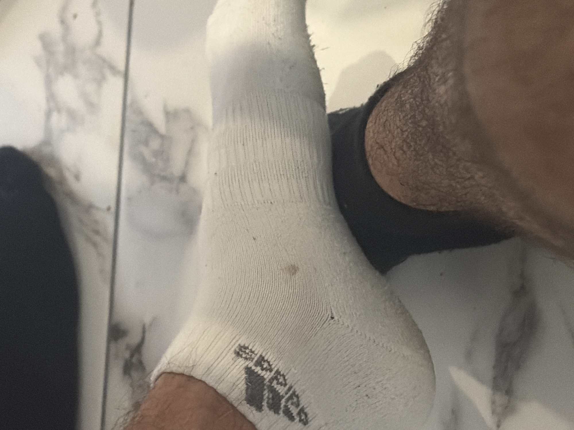 The image shows a close-up view of two feet. One foot is wearing a white athletic sock with the Adidas logo, and the other foot is wearing a dark sock. The feet are resting on a tiled floor. The scene is indoors. The image does not provide any information about the city of Podgumer, Bulgaria.