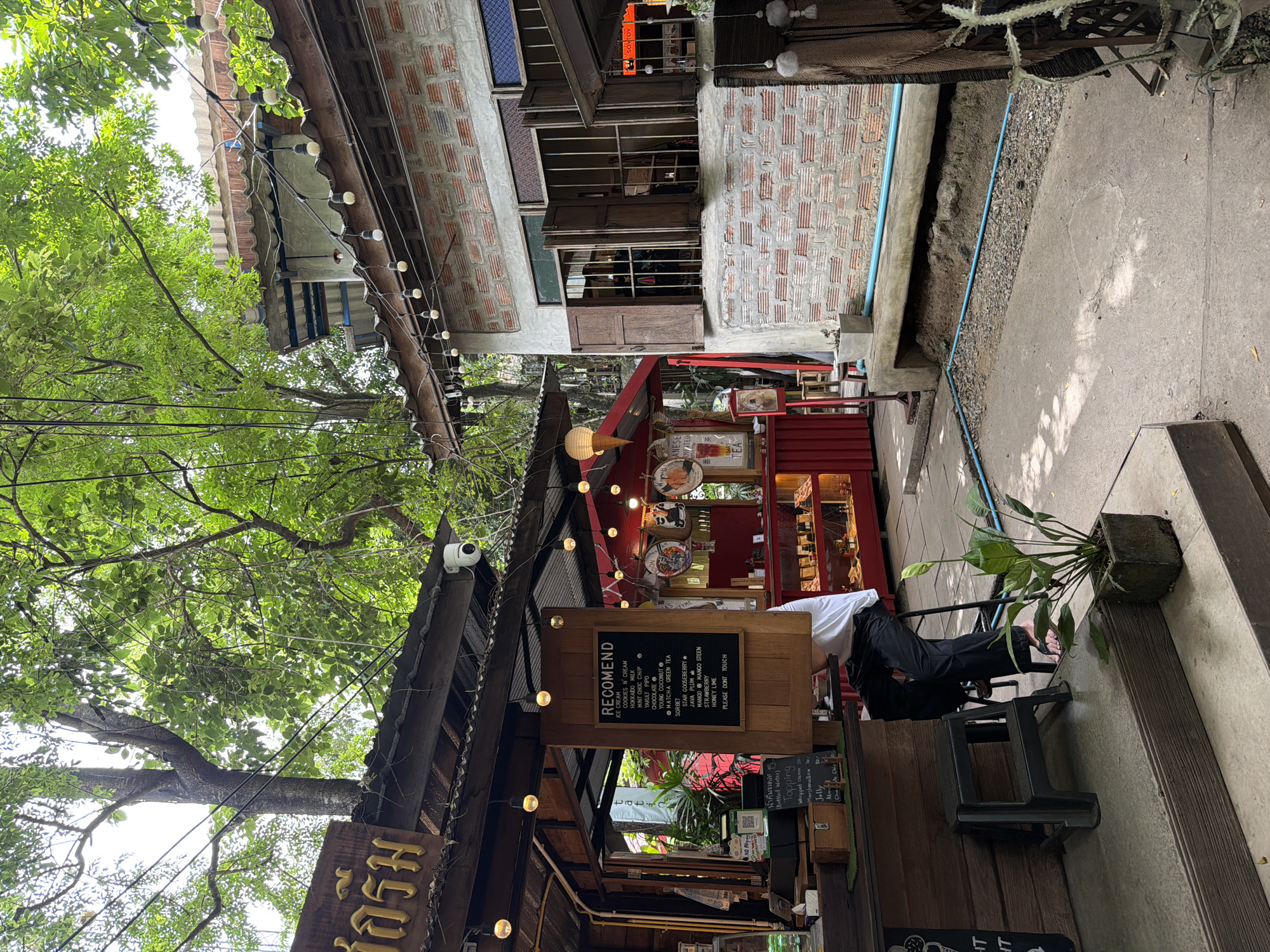 The image shows an outdoor seating area of a cafe in Ban Sai Kham, Thailand. In the foreground, a person is seated on a stool at a wooden counter. To the left, a menu board displays recommendations for ice cream and other desserts. Further back, a red food stall is visible with illuminated displays of products. The cafe is surrounded by trees and foliage, with a rustic, open-air design. String lights are strung across the scene, and traditional brick architecture is present in the background. The overall ambiance suggests a casual and charming setting for enjoying food and drinks.