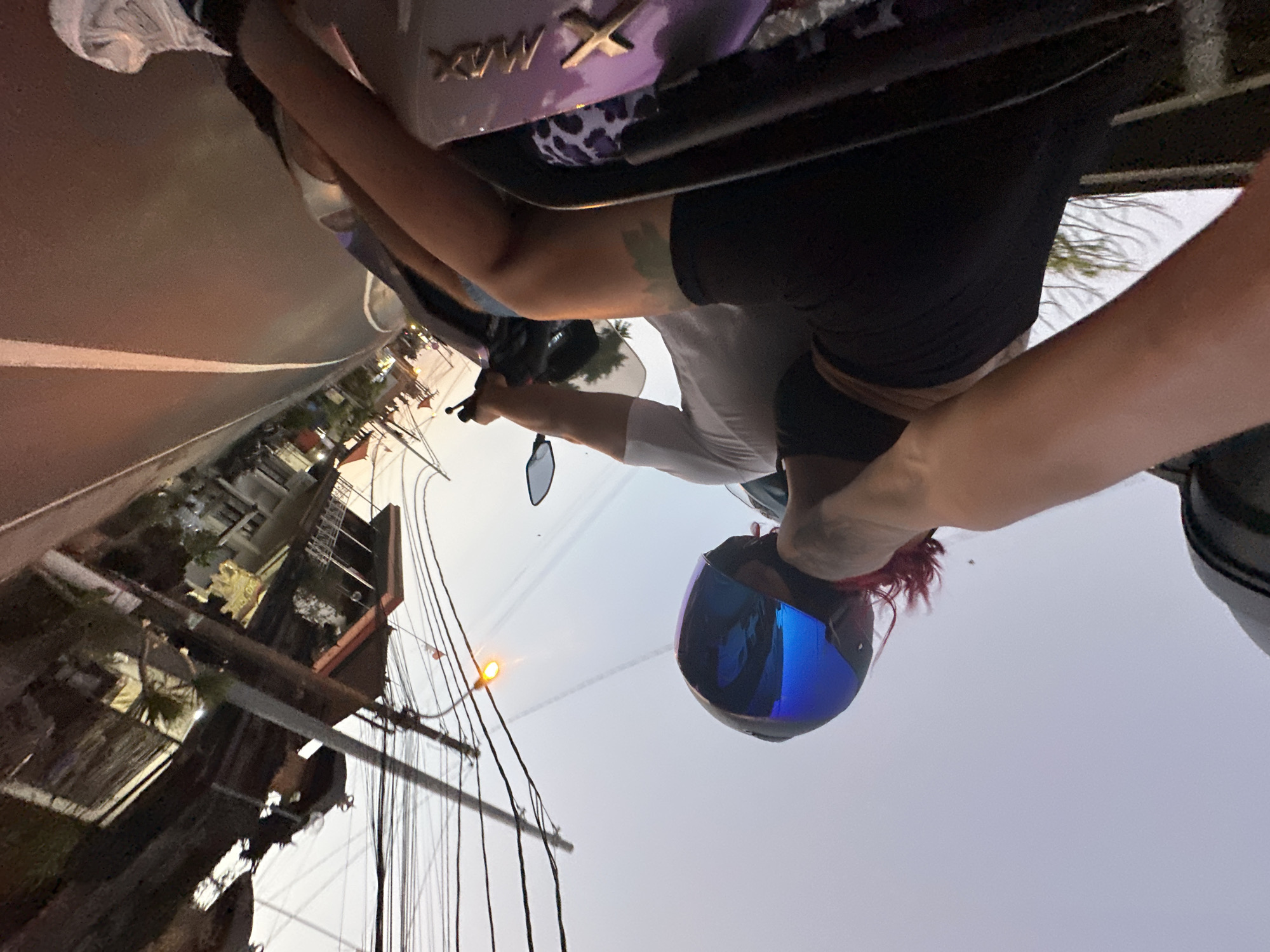 A woman wearing a blue-tinted motorcycle helmet is riding a motorcycle on a road in North Kuta, Indonesia. The road runs alongside buildings with electrical wires running overhead. The woman is wearing a black shirt and black shorts. The motorcycle is purple.