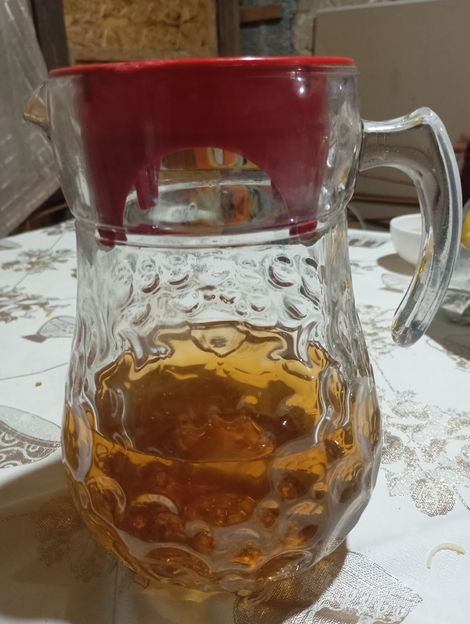 A glass pitcher with a red lid is filled with amber liquid and sits on a white tablecloth with a floral pattern. The pitcher has a textured surface. The background shows a wooden wall and a grey concrete wall. The scene is indoors and appears to be in a residential setting in the city of Qurama, Uzbekistan. There are no people visible in the image.