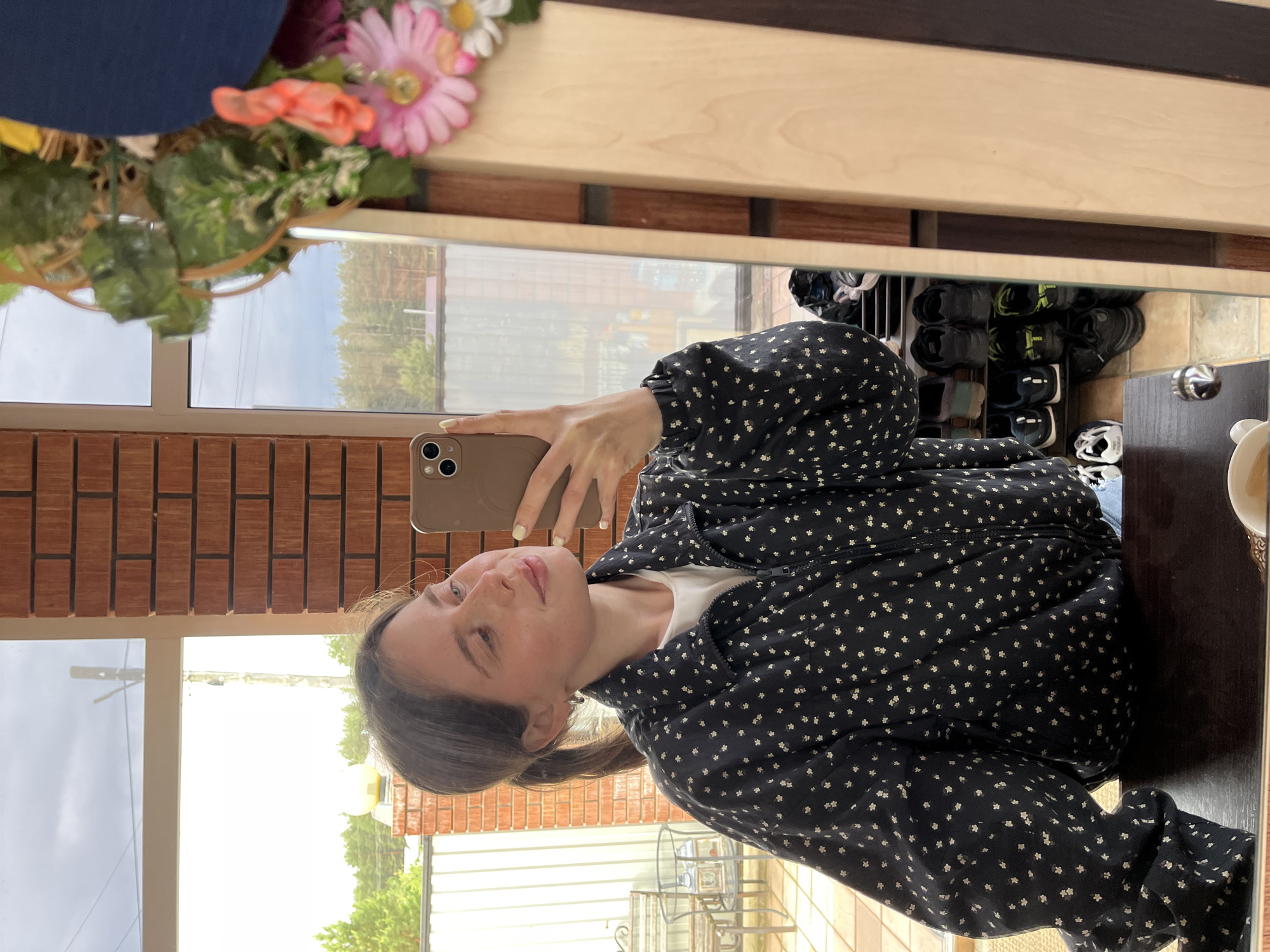 A woman is taking a selfie in a mirror. She is wearing a black jacket with small white floral patterns over a white shirt. Her hair is pulled back in a low ponytail. The mirror is framed with light wood and a dark wood trim. In the reflection, a glimpse of the room shows a shelf of shoes, a cup, a red brick wall, and outdoor furniture. A garland of flowers is attached to the mirror frame. Based on the environment in the reflection, specifically the brick wall style and outdoor furniture, it's plausible this photo was taken in Chaikovsky, Russia, although confirmation is needed to confirm the exact location.