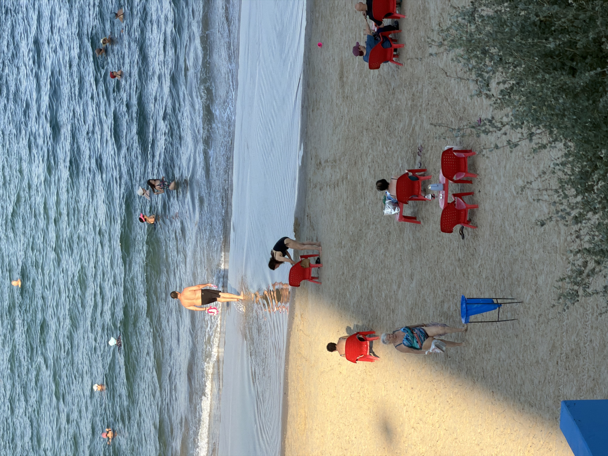 The image depicts a crowded beach scene in Bat Yam, Israel. The sea is calm with small waves, and several people are swimming and wading in the water. On the sandy shore, there are numerous beach chairs and tables set up, occupied by sunbathers and groups of people. A man in black shorts and a bare chest walks from the sea onto the beach. Another woman in a black swimsuit is standing near a red chair. A woman in a colorful swimsuit is walking across the sand. In the background, a blue trash can is visible. The overall scene is one of a typical summer day at a popular beach destination.