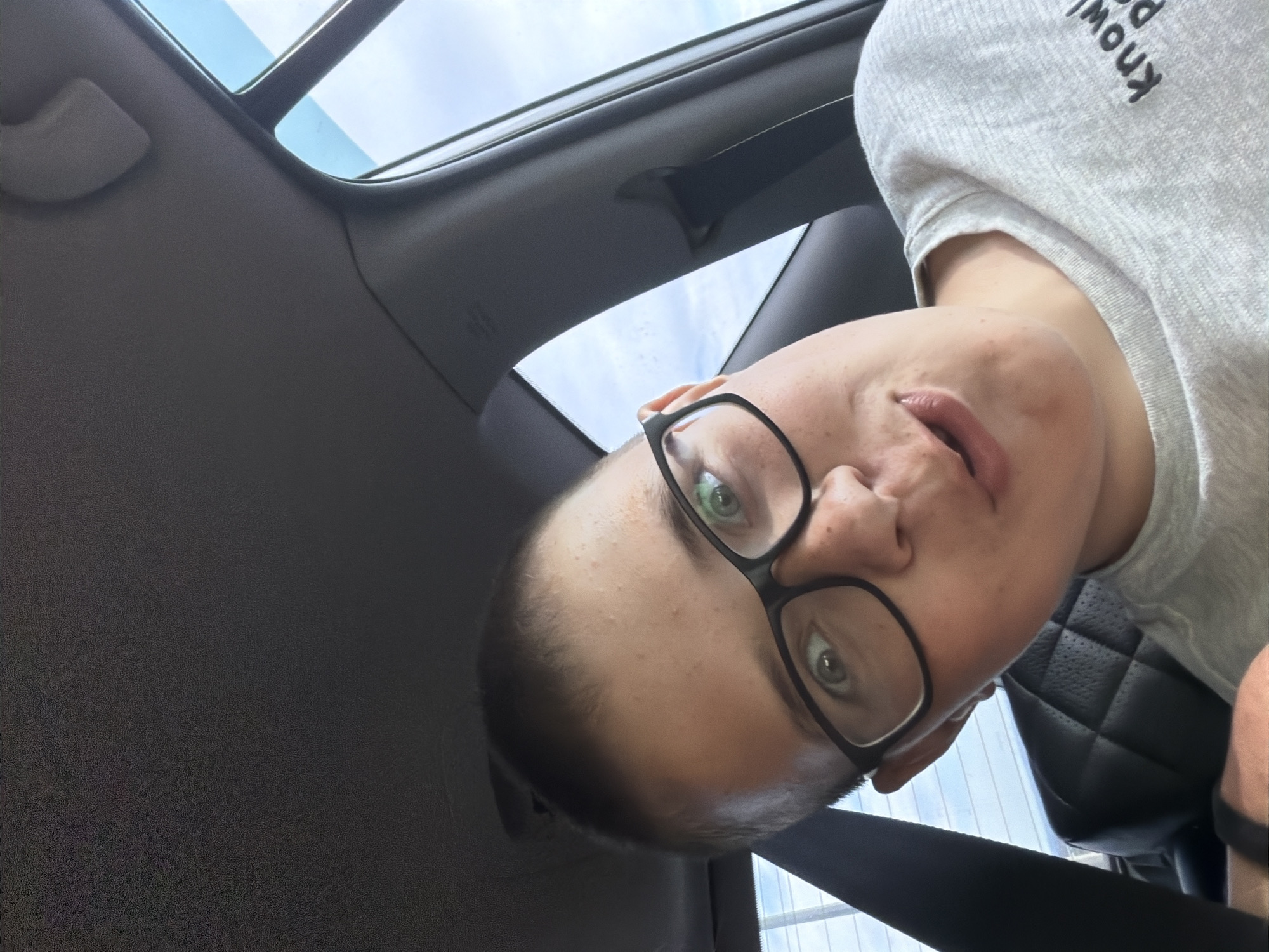 A close-up, slightly tilted selfie is captured from inside a car. The subject, a young person with short, dark hair and glasses, is looking directly at the camera with wide eyes and a slightly open mouth. They are wearing a grey t-shirt with some text on it that is partially visible. The background shows the interior of the car, including a dark leather seat and the car's headliner. Outside the window, a bright sky with some clouds is visible. The overall impression is that of a casual, spontaneous photograph taken while traveling. The image does not contain any elements that can indicate its location in the city of Новое Маврино, Russia.