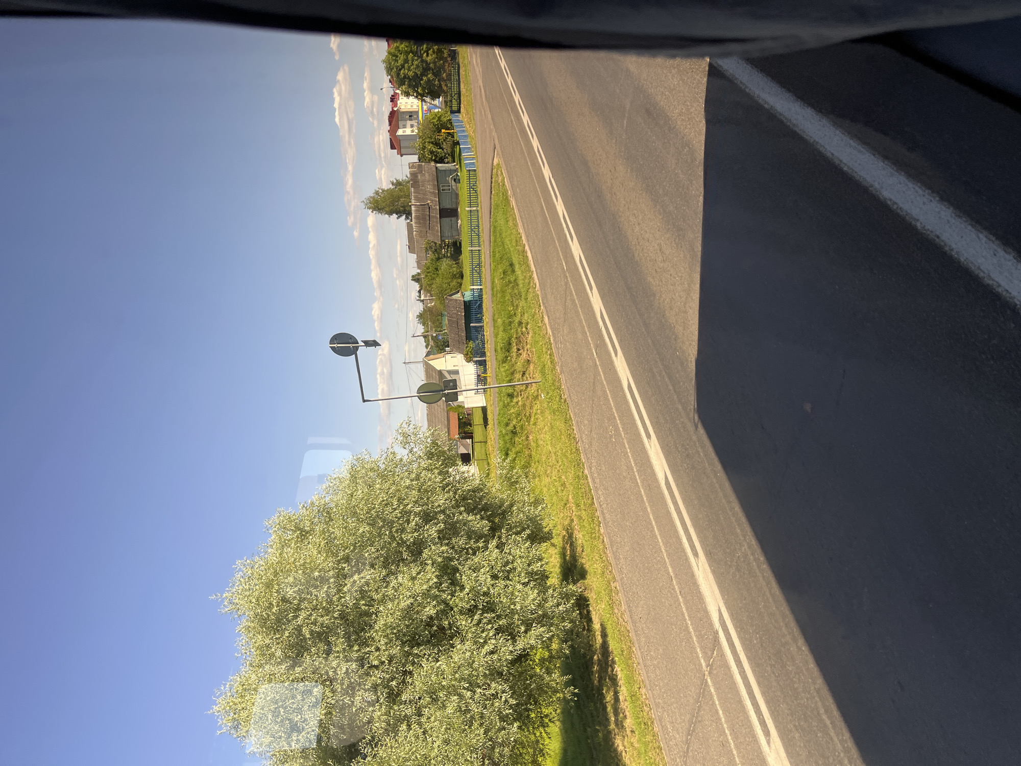 This is a medium-shot, eye-level, horizontal, outdoor, daytime, photographic image taken from a vehicle, likely a bus, looking out of a window, showing a view of a rural road with scattered houses and trees under a clear blue sky with scattered clouds. The road is asphalt with double white lines in the center and a white line on the right edge. There is a large, leafy green tree on the left side of the road, casting a shadow on the road. The right side of the image is dominated by the dark frame of the vehicle window. Across the road, there are several houses, some with blue fences, and more trees. Two traffic signs are visible on poles. The scene depicts a semi-urban or rural environment.