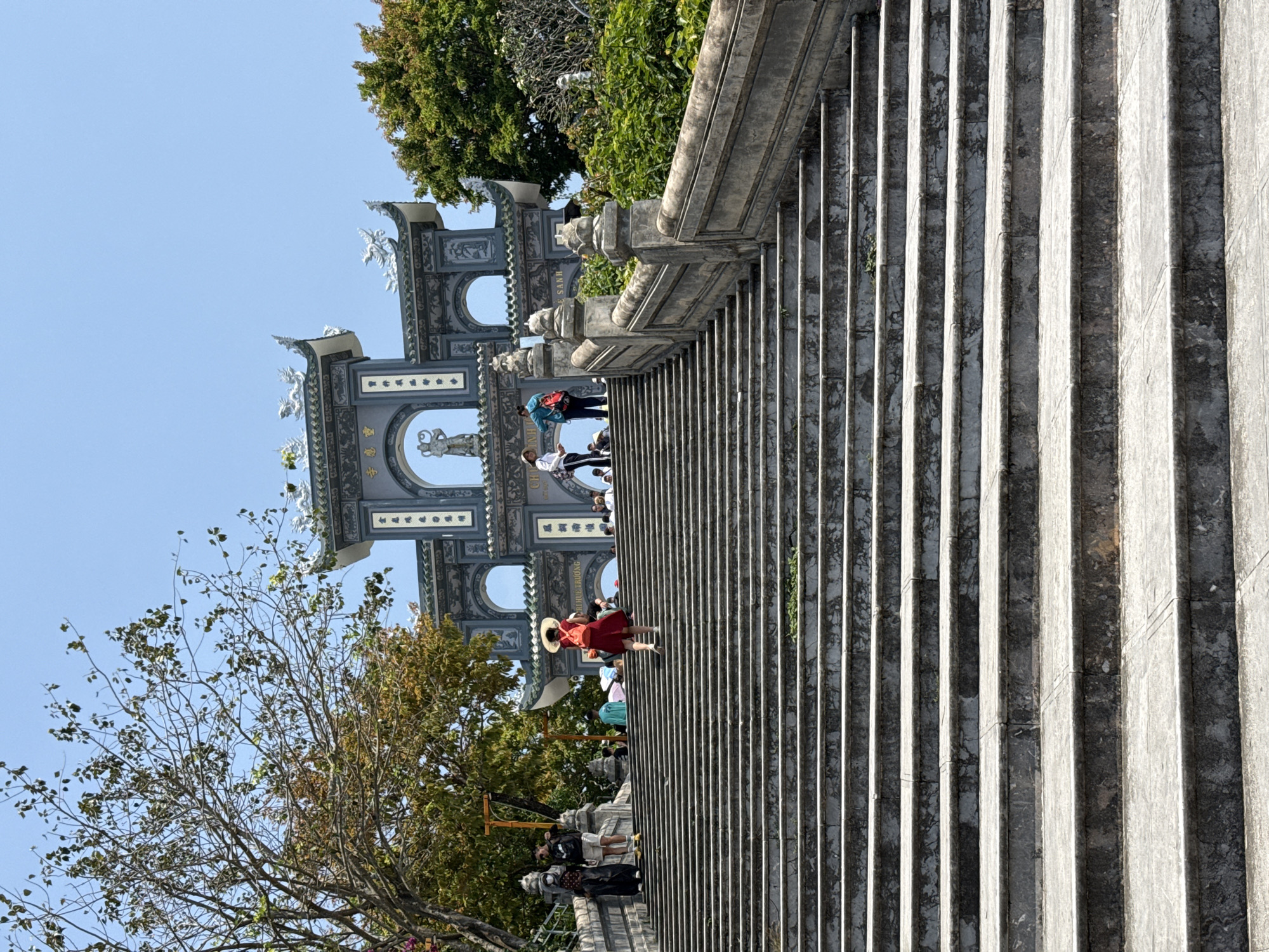 A wide, multi-tiered grey stone staircase ascends towards a triple-arched ornate gateway, identified as the entrance to Linh Ung Pagoda on Son Tra Peninsula in Đà Nẵng, Vietnam. The staircase exhibits signs of wear and sparse vegetation between steps. Approximately ten individuals are visible on the stairs and near the gate. A person wearing a red dress, a wide-brimmed straw hat, and an orange backpack is centrally positioned, ascending the stairs with their back to the viewer. To the left, two individuals stand near a stone balustrade; one wears a dark outfit and face mask, the other a black t-shirt and light shorts. The gateway is dark grey with teal-colored roofs, featuring intricate white sculptural elements including mythological creatures and human figures within its arches. Vietnamese script, including 'CHÙA LINH ỨNG', is visible on its pillars. Green foliage from various trees frames the scene on both sides, with a clear blue sky overhead.