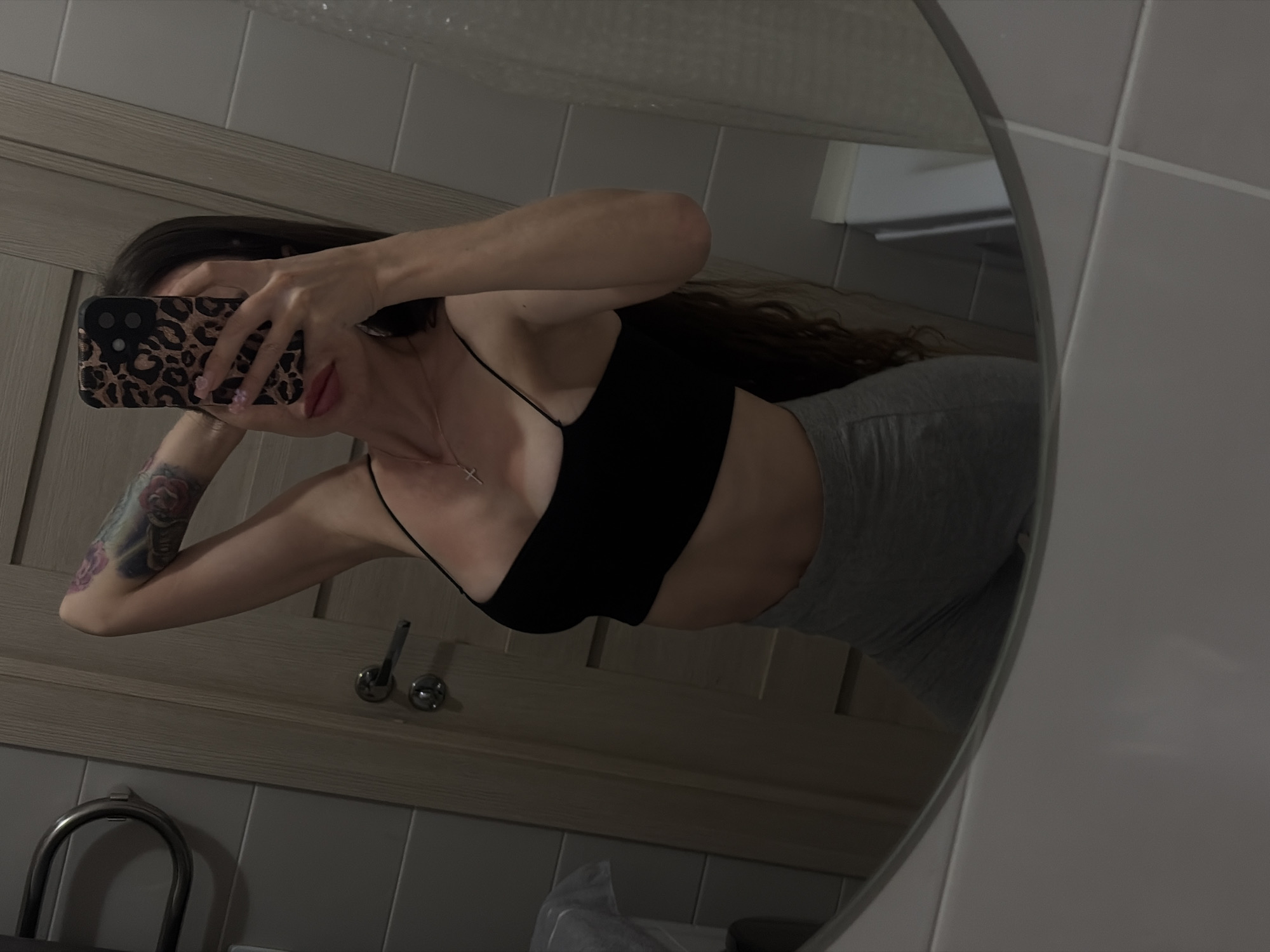 The image is a mirror selfie taken in a bathroom. A woman with long, dark hair and a tattoo on her left arm is holding her phone to take the photo. She is wearing a black tank top and grey leggings. The bathroom has tiled walls and a wooden door.