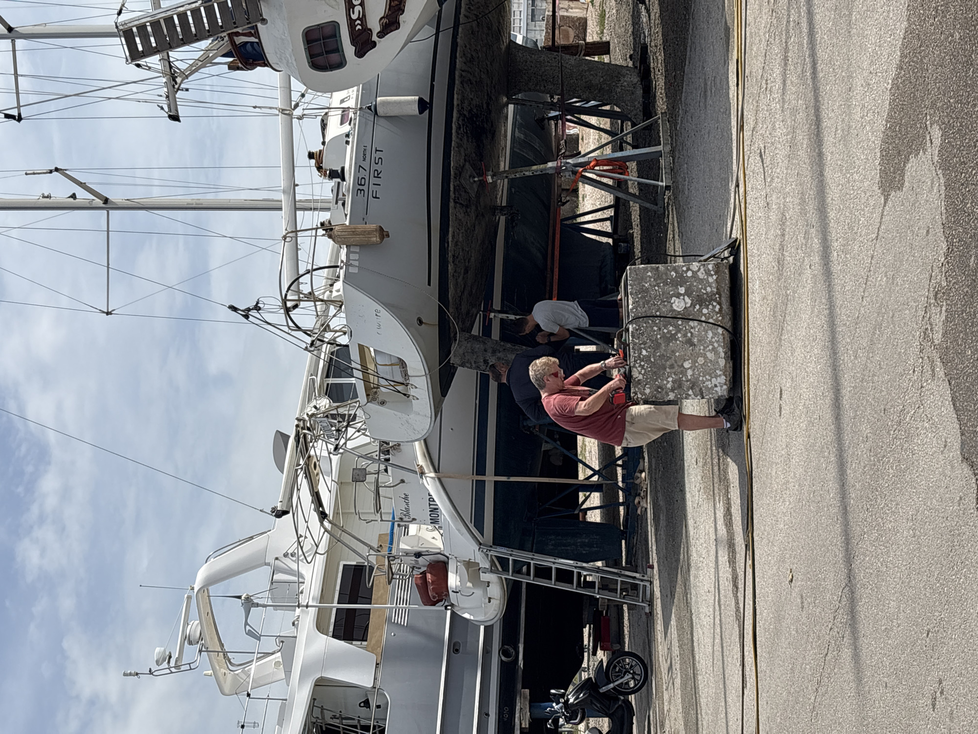 The image captures an outdoor scene at a boatyard or dry dock in Bar, Montenegro, under a bright, partly cloudy sky. Several boats, predominantly sailboats and one motor yacht, are visible, lifted out of the water and resting on metal support stands on a concrete surface.
In the foreground, a man wearing a red t-shirt, khaki shorts, and sunglasses is actively working next to a large, weathered concrete block, possibly operating a tool or engaging with some equipment. Another man in a grey t-shirt is partially visible further back, near the hull of one of the sailboats, also seemingly involved in maintenance.
Notable details on the boats include the text "36.7 FIRST" prominently displayed on the hull of a sailboat, with "NORTH E" printed in smaller letters below it. Another boat in the lower-left section features "Blanche" and partially obscured "MONTR" (likely part of a larger word like Montpellier) on its stern. The boats are equipped with rigging, masts, and ladders for access. Fenders are hanging from the sides of some vessels.
Other objects include a motorcycle or scooter parked near the boats, and various cables or hoses lying on the concrete ground. The ground itself is textured and shows signs of use typical of a working dock area. Long shadows cast by the boats and other structures indicate it is daytime. The scene depicts ongoing maintenance and preparation of boats in a maritime setting.