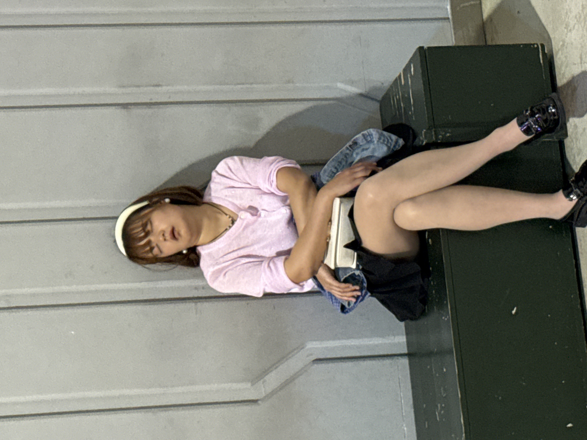 A woman is lying on an object that appears to be dark-green metal crates stacked in a corner of a room. She is wearing a light pink shirt, a headband, and black shoes with silver buckles. Her eyes are closed, and her legs are extended on the crates. A jean jacket and a black skirt or shorts are around her. The wall behind her is a light gray color. This photograph was taken in 江北城街道, China.