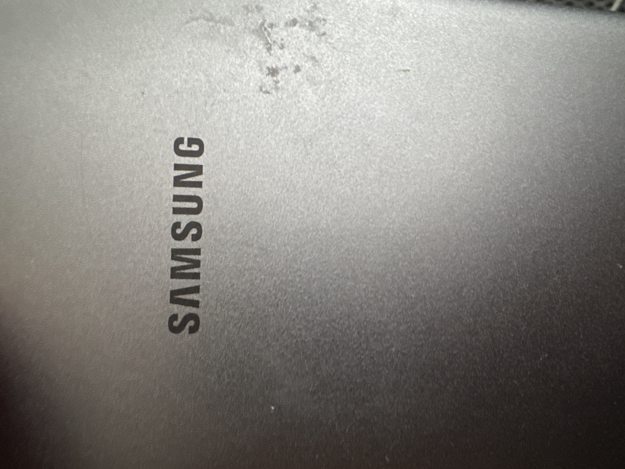 The image shows the SAMSUNG logo embossed on a dark gray, textured surface. The logo is centrally located and appears to be made of a darker material than the surrounding surface. The surface itself has a subtle brushed metal texture and exhibits some light reflectivity. There are a few small, light-colored specks or blemishes on the surface, particularly to the right of the logo. The lighting appears to be coming from the upper right, creating a slight gradient of brightness across the image, with the upper right being lighter. No specific location in Zaokskiy, Russia, or any environmental context beyond the object itself is discernible. The image is a close-up shot of the product branding.