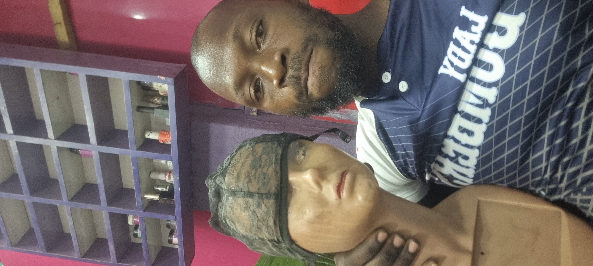 A man with a close-cropped haircut and a beard stands next to a mannequin head. The man's shirt is navy blue with a white and navy diamond pattern and white text that is partially obscured. The mannequin head wears a black lace cap, and the man's hand is visible on the mannequin's neck. To the left, there's a purple shelf with various items on it. The room has a pink wall. There is no concrete evidence within the image to confirm its location in Lagos, Nigeria. Further information would be needed to confirm the location.