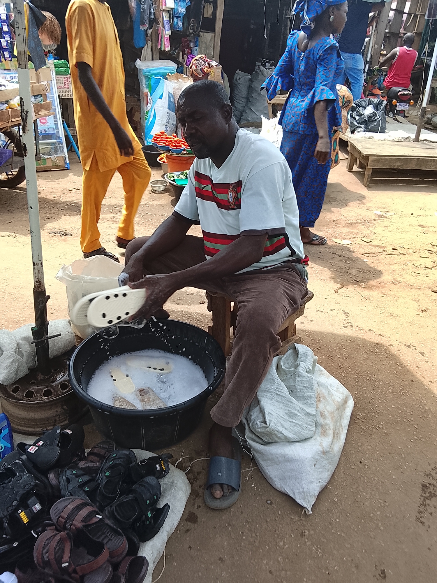 In Akwanga, Nigeria, a man sits on a wooden stool, cleaning white sandal soles in a black bucket of soapy water. He wears a white t-shirt with red and green stripes, brown pants, and blue sandals. Beside him is a white sack, and a pile of sandals is to his left. He is positioned near a metal pole. In the background are other people, some in traditional clothing, and structures typical of an outdoor market.