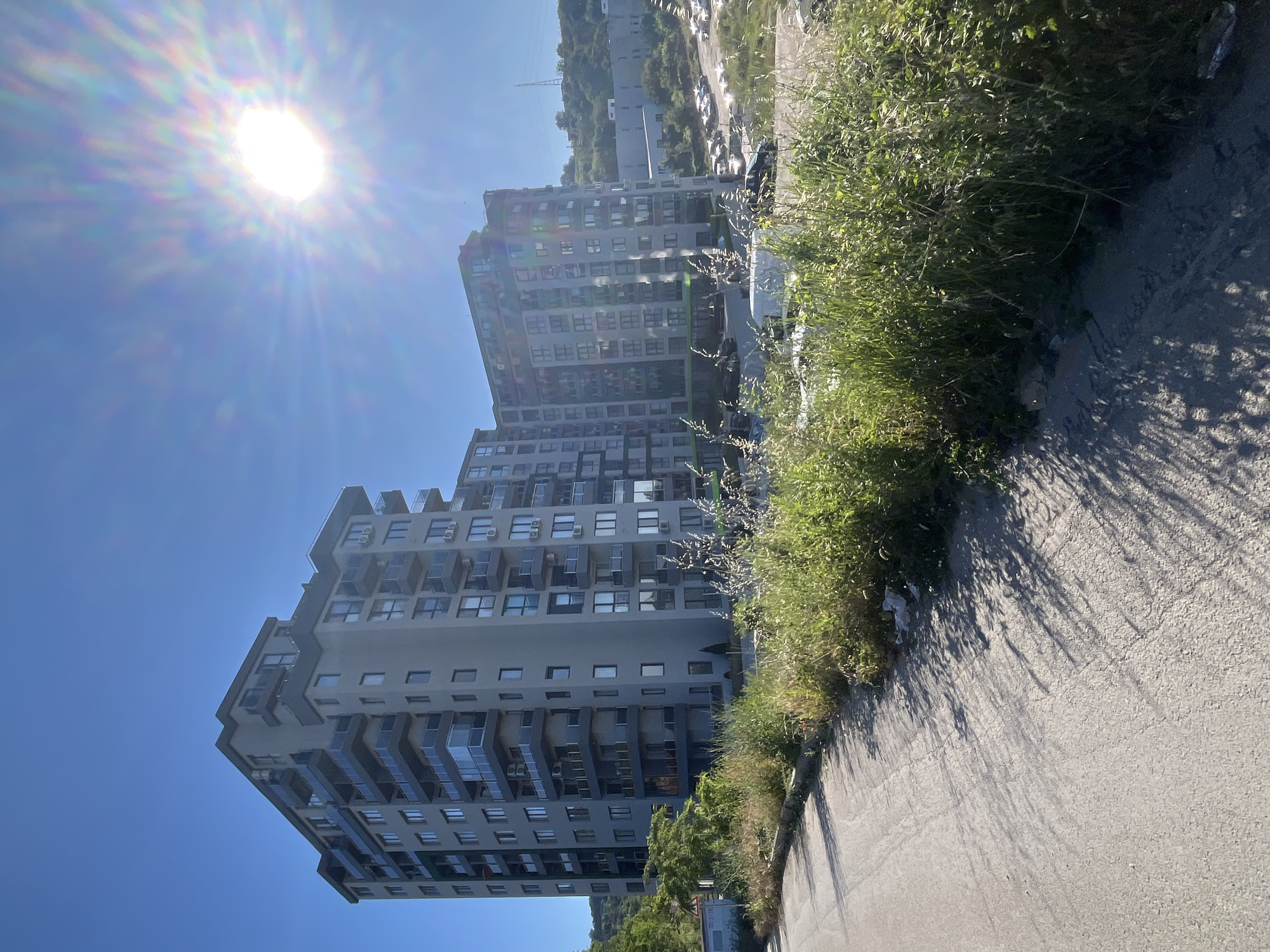 The image depicts a large, modern apartment building with multiple stories and balconies. The building is light gray in color and has numerous windows. It is situated on a hillside overlooking a parking lot filled with cars. The foreground shows a paved road and a patch of overgrown grass and weeds. The sky is a clear, bright blue with the sun prominently visible in the upper right corner, casting lens flares and rays. The scene appears to be in Varna, Bulgaria, given the architectural style and the general setting which aligns with descriptions of that city. There are no people visible in the image. The overall impression is that of an urban or suburban residential area on a sunny day.