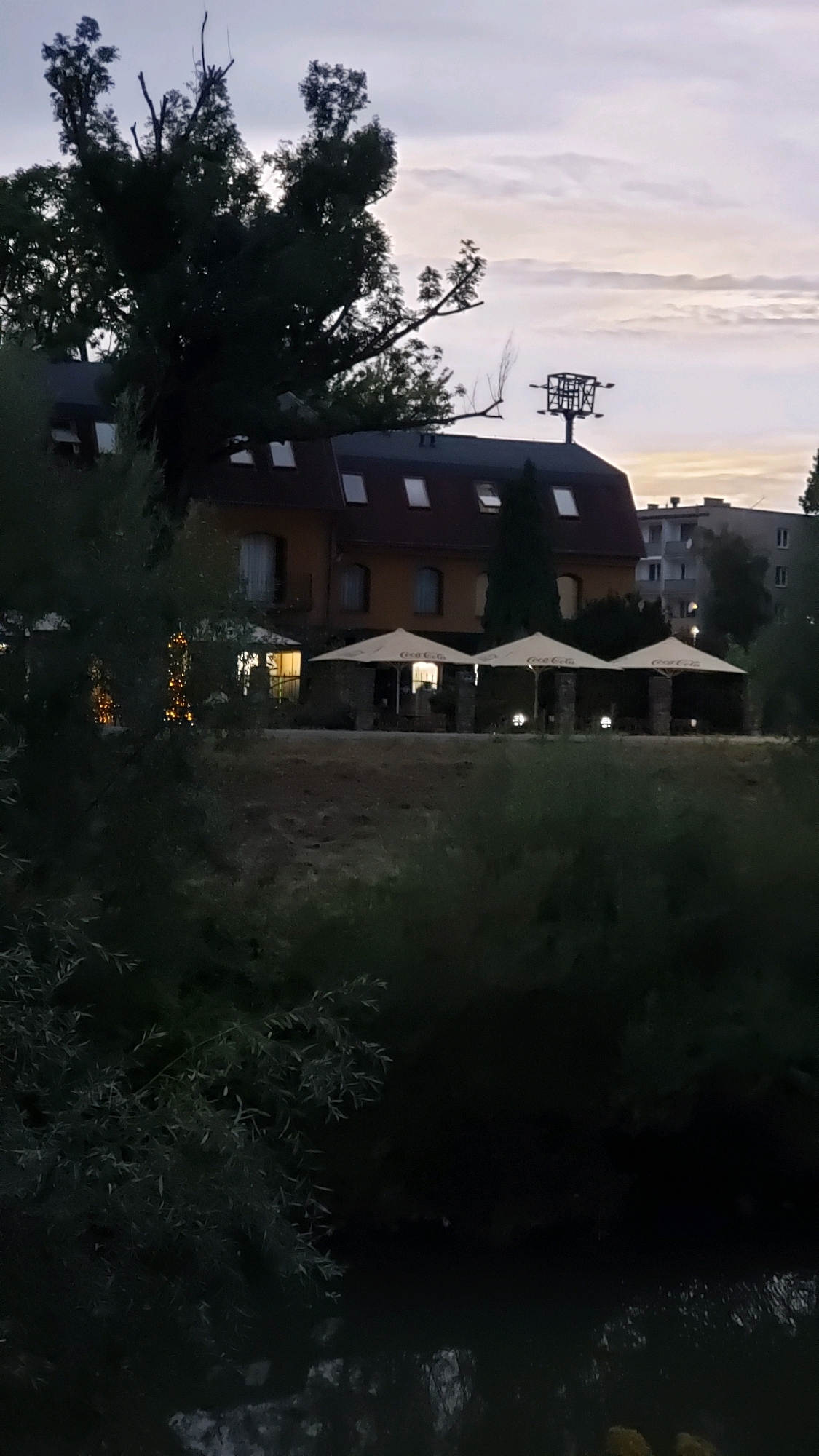 The image depicts the exterior of a building that appears to be a restaurant or hotel, situated along a body of water. The building has an orange facade, a dark-colored roof with multiple skylights, and several arched windows on the first floor. In front of the building, there is an outdoor seating area with three large umbrellas, each with the "Coca-Cola" logo visible. Stone pillars support the umbrellas, and there are lights illuminating the seating area. String lights are also visible on the left side of the building, suggesting a festive atmosphere.
Behind the main building, there are trees and a building in the background, which seems to be an apartment complex. The sky is overcast with a gradient of colors suggesting either dawn or dusk. In the foreground, there is dense foliage obscuring the view of the water. A significant tree with bare branches and dark foliage is prominent on the left side of the image, partially covering the building. A distinctive structure, resembling a metal frame with multiple levels, is visible above the trees and building.
Based on the presence of the "Coca-Cola" branded umbrellas and the architectural style, this scene is likely the restaurant "Stella bistro" located at Sládkovičova 43, Prešov, Slovakia. The body of water in the foreground is the Torysa River.