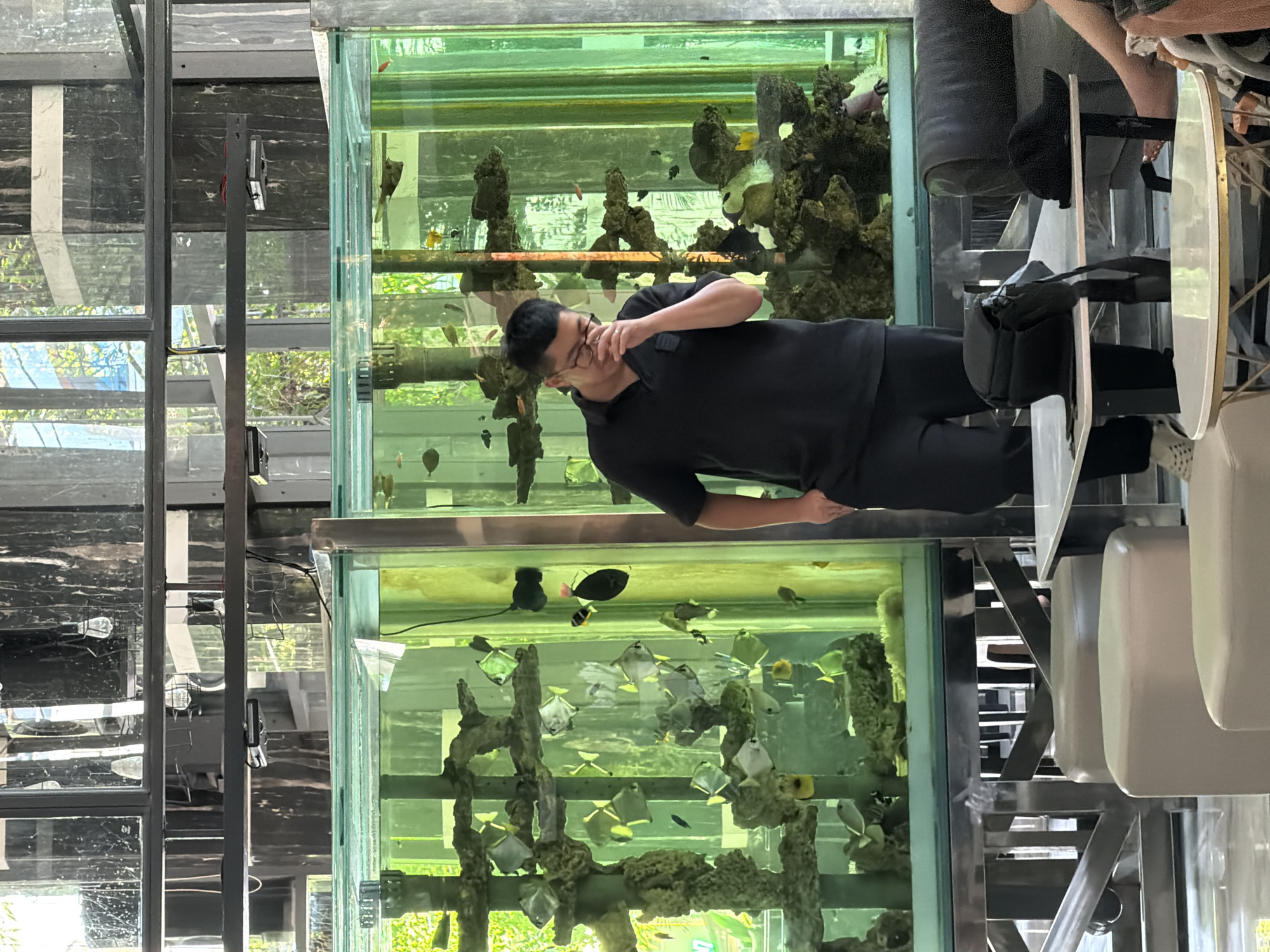 A man wearing glasses and a black polo shirt stands in front of a large aquarium filled with fish and coral. The aquarium is part of a modern interior space with large windows and reflective surfaces. The man is holding his hand to his face, and appears to be looking down. Several round, upholstered ottomans are visible in the foreground, along with a table with a black bag and a black cap on it. The scene suggests a public indoor space, possibly a cafe or lobby, in Nha Trang, Vietnam.
