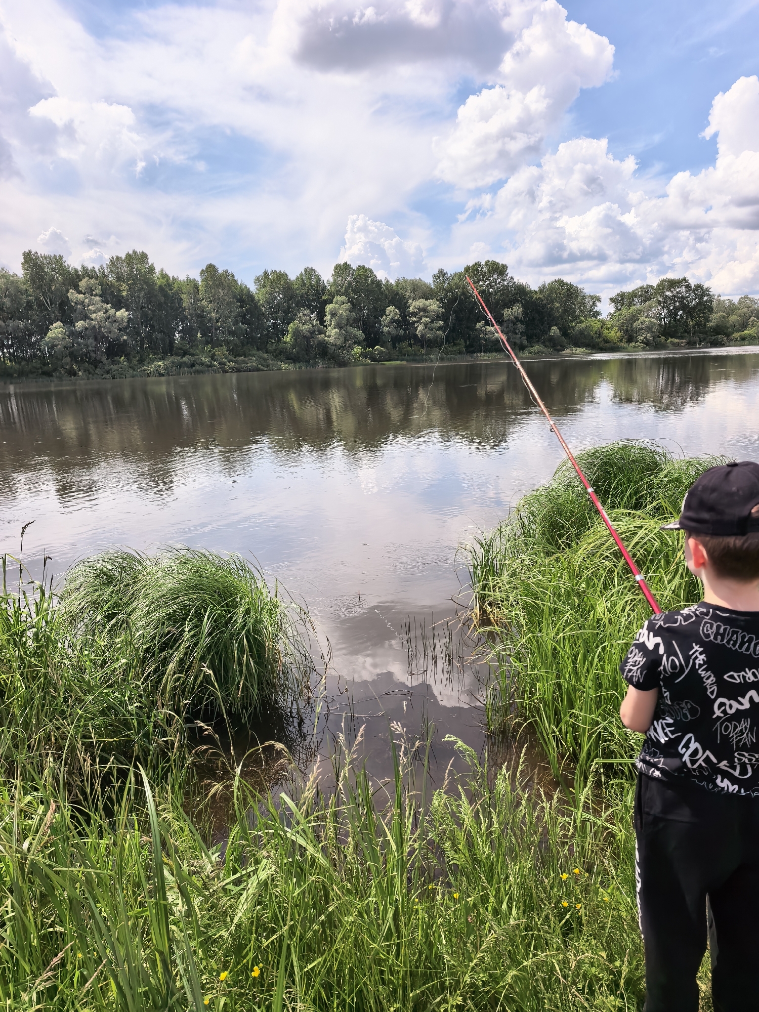 A young boy, seen from behind, stands on the grassy bank of a wide, calm river, holding a red fishing rod. He is wearing a black t-shirt with a white graphic print and a black baseball cap. Tall green reeds and grass grow along the river's edge, partially obscuring the water's surface. Across the river, a dense line of green trees forms the opposite bank, with a reflection of the trees and the cloudy sky visible on the water. The sky is bright blue with scattered white cumulus clouds. The scene appears to be set in a natural, possibly rural or park-like area, given the river and tree line. The location is identified as Kuzminovka, Russia.