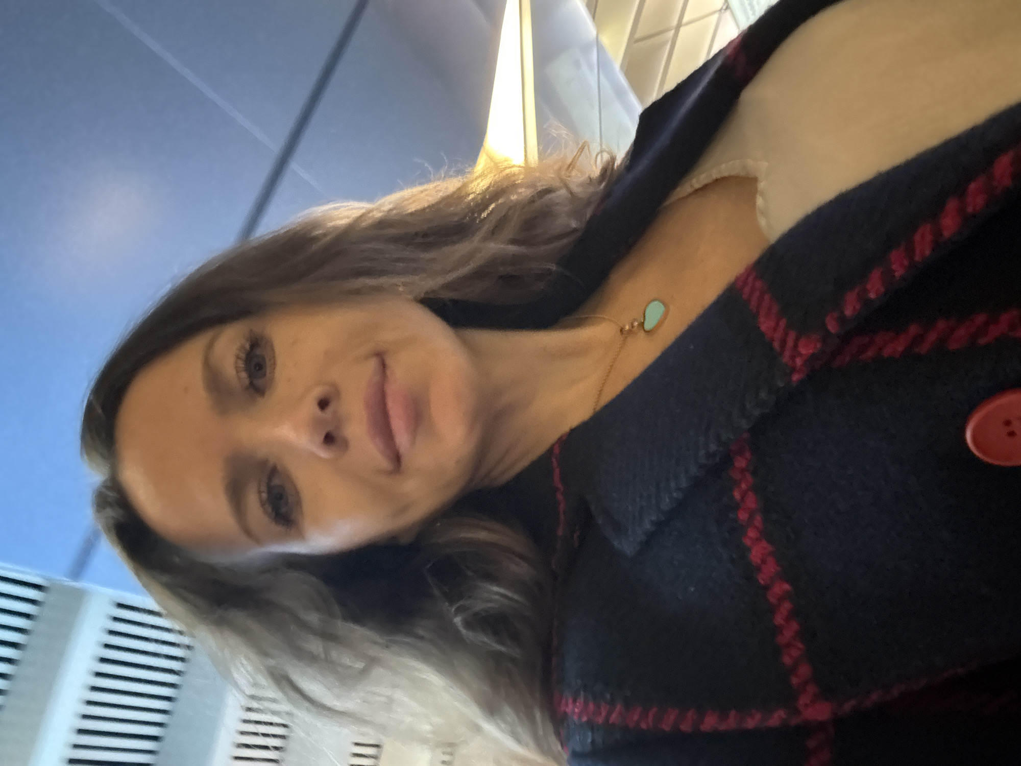 A woman is taking a selfie. She has long, blonde hair and is wearing a navy blue coat with a red plaid pattern, and a white shirt underneath. She is also wearing a necklace with a teal heart-shaped pendant. The background appears to be an indoor setting with fluorescent lighting, possibly a public building or transportation hub. The angle of the photo suggests the woman is looking up at the camera, and her expression is neutral. There are no visible signs of the time of day, weather, or specific emotions. The location context is Moscow, Russia, but there are no specific visual cues in the image to confirm this.