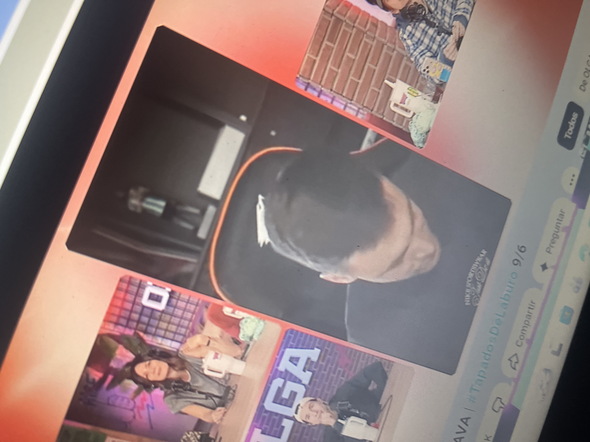 The image is a close-up, angled photograph of a computer screen displaying a video stream. Multiple individuals are visible in separate video feeds on the screen.
In the top left feed, a woman with long dark hair and headphones is seated at a desk, holding a white mug. Behind her, a stylized graphic with pink and purple neon elements is visible. A white square with a black "O" is on the wall behind her.
In the bottom left feed, a person with a cap is seated at a desk, holding a white mug. A large purple graphic with white letters spelling "LGA" is behind them.
The largest feed in the center shows the back of a person's head, with dark hair, looking down. This feed appears to be a close-up of a person in a chair. A portion of a gaming chair with red trim is visible.
To the right of the central feed, a brick wall texture is visible. In this section, there are more video feeds. One shows a person wearing a blue and white plaid shirt, holding a mobile phone. There are objects on a shelf, including a white pitcher-like container and a blue and yellow item.
At the bottom of the screen, there are interface elements for a video platform, including text that reads "AVA | #TapadosDeLaburo 9/6", "Compartir" (Share), and "Todos" (All). There is also a visible portion of text that appears to read "De OLGA".
There is no information within the image that specifies a location in Santa Ana, Venezuela.