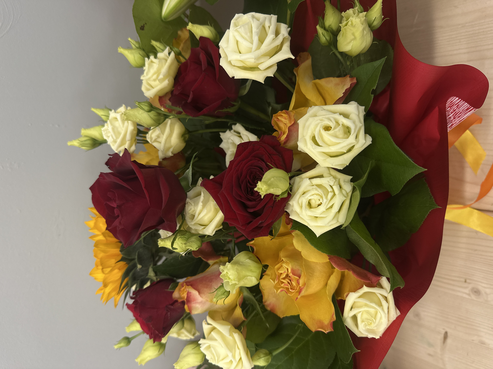 A bouquet of flowers, including red and white roses, yellow flowers resembling sunflowers, and green buds, is presented in red wrapping paper. A yellow ribbon is attached to the bouquet. The background appears to be a gray wall on one side and a light-colored wooden surface on the other. The city of Riga, Latvia, is not ascertainable from the image. No people are visible.