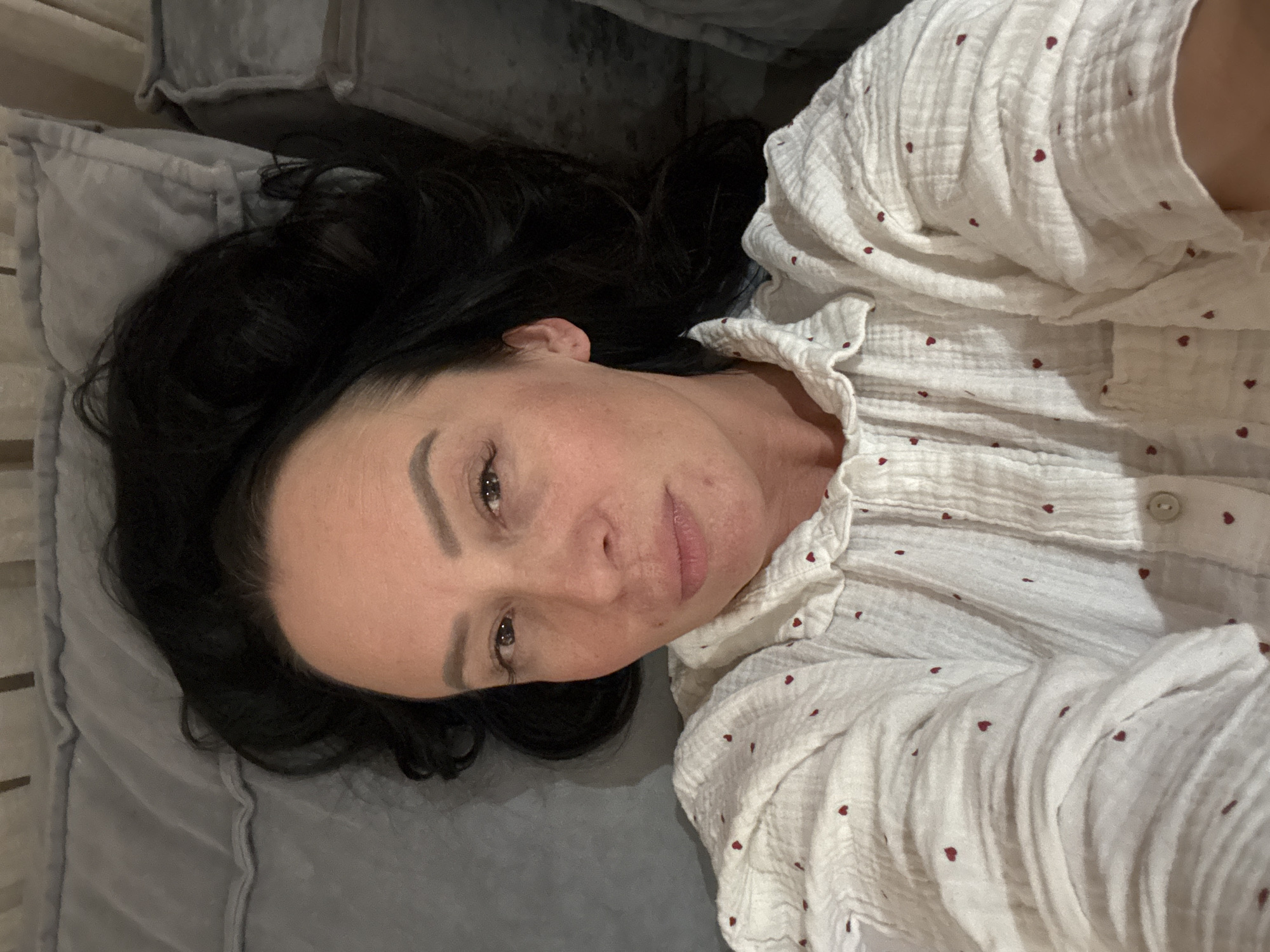 A female individual is depicted in a close-up, overhead shot, lying supine on a bed or cushioned surface. Her dark, wavy hair is spread out behind her head on a light gray quilted pillow. Her face, with fair skin, dark eyebrows, and dark eyes, is directed towards the camera with a neutral expression. She is wearing a white, textured, collared shirt adorned with small red heart patterns, featuring a visible buttoned placket. The immediate background consists of further light gray upholstered elements, possibly additional pillows or a headboard. The scene is located in the city of Chaikovsky, Russia.