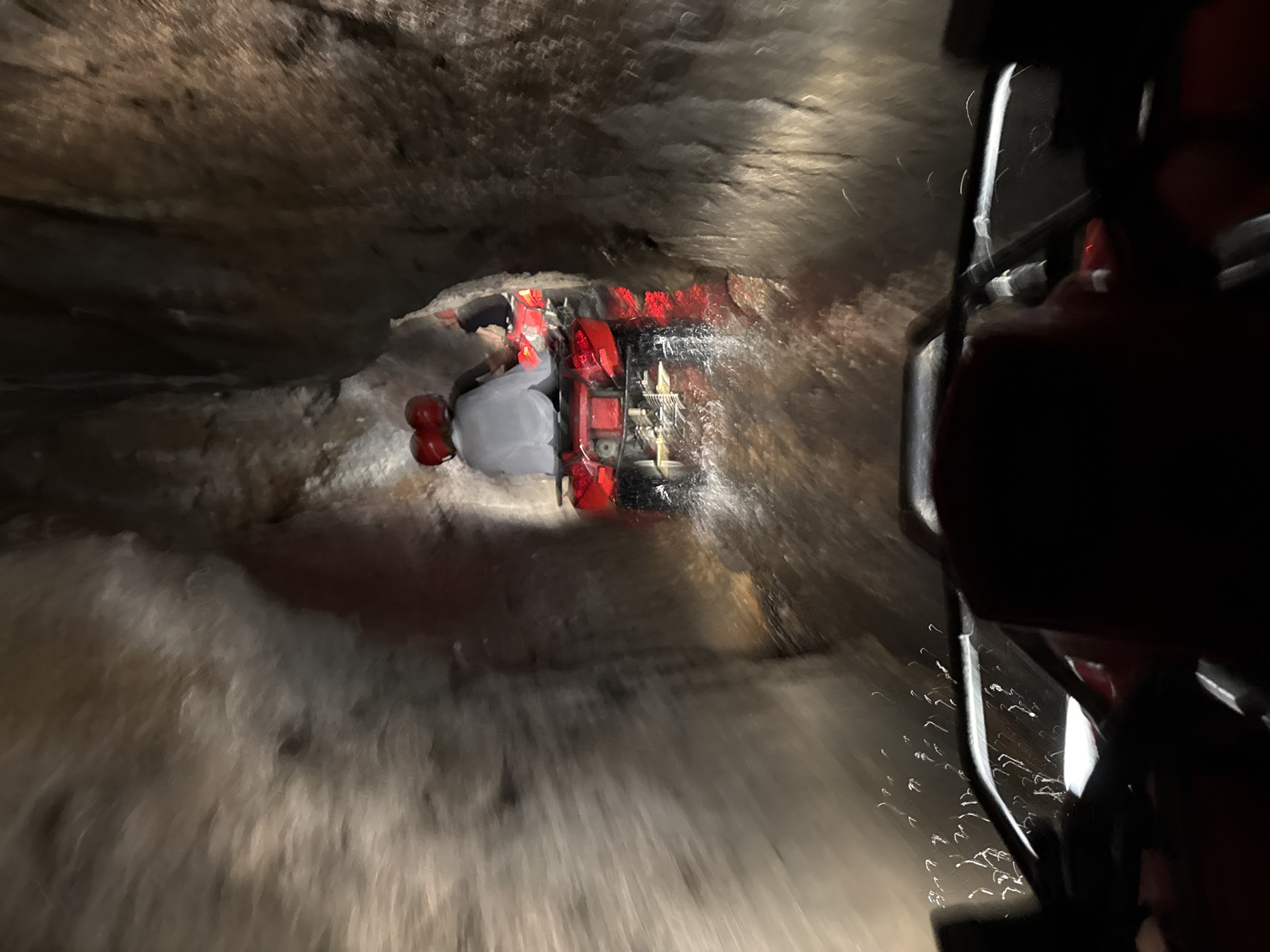 The image captures a first-person perspective from an All-Terrain Vehicle (ATV) traveling through a dark, narrow, and wet natural cave or tunnel in Payangan, Indonesia. In the immediate foreground, the dark handlebars and a red section of the viewer's ATV are visible, with streaks of water or mud indicating motion and a wet environment. Directly ahead, another red ATV is observed from the rear, carrying two individuals. Both riders are wearing red helmets. The lead rider is clothed in a dark top, while the passenger wears a light-colored, possibly white, long-sleeved top. The ATV's red tail lights are illuminated, and water is actively splashing around its rear wheels, suggesting movement through standing water or mud. The walls of the cave or tunnel consist of rough, uneven rock formations, appearing damp or wet, with some areas exhibiting light reflections. The environment is dimly lit, consistent with an underground setting.