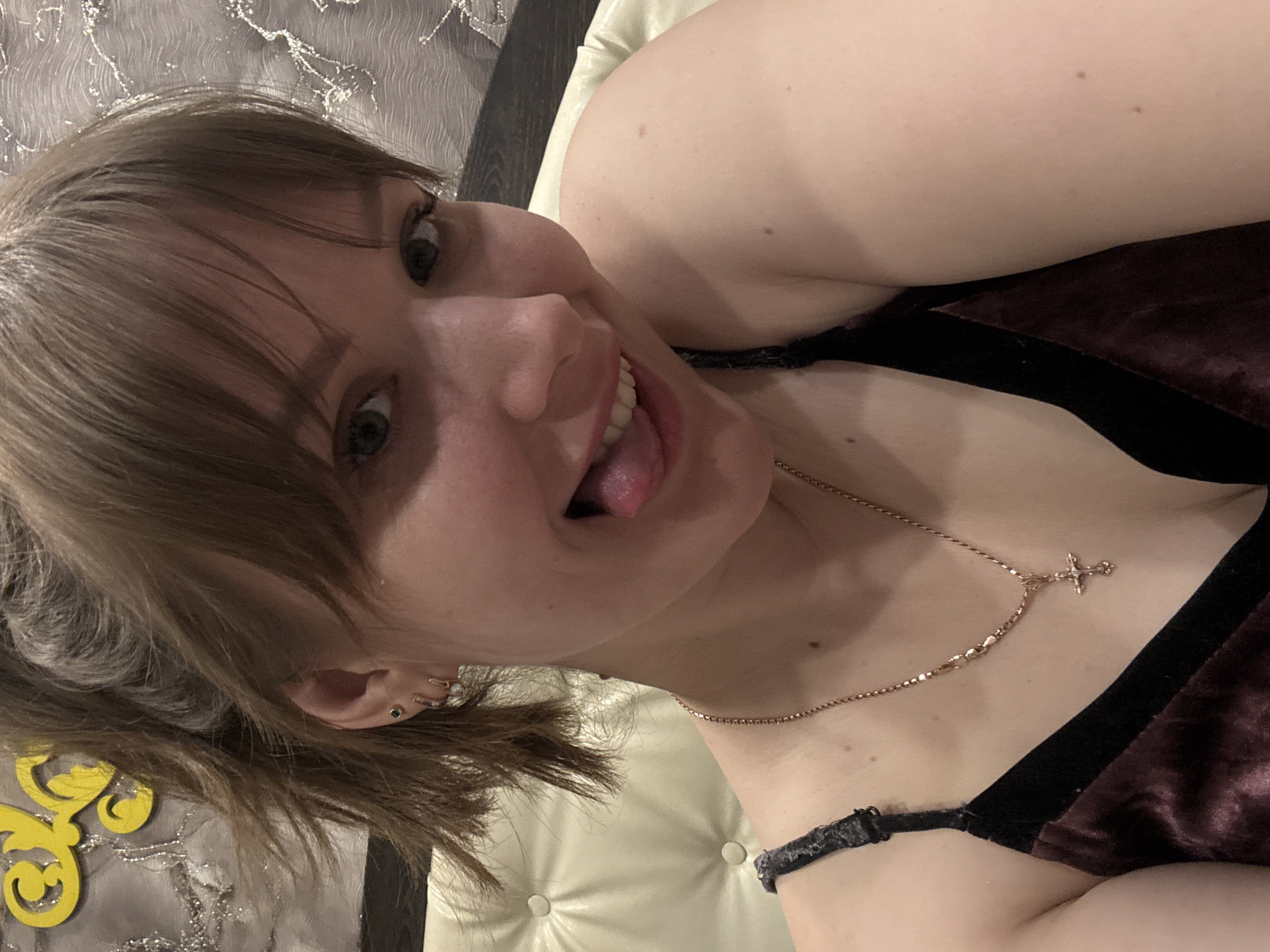 A light-skinned woman with shoulder-length brown hair is seen from a close-up, slightly angled perspective. She is sticking her tongue out and smiling. She is wearing a dark-colored velvet garment with black straps and a gold necklace with a cross pendant. She has multiple piercings in her left ear. The background includes a tufted white headboard and a patterned fabric. The location, according to the prompt, is Chaikovsky, Russia, but this cannot be verified from the image itself.