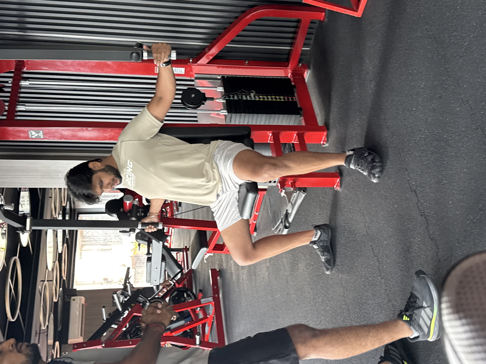 In a gym setting in Kolkata, India, a man is seated on a weight machine, performing a chest fly exercise. He is wearing a light-colored t-shirt with a graphic print and white shorts. His arms are extended outwards, holding the handles of the machine. Another person, partially visible on the left side of the frame, is standing and appears to be observing or assisting. The gym features red and black equipment, with a black striped wall in the background. The floor is dark grey and textured. Other gym equipment, including weight racks and benches, are visible in the background.