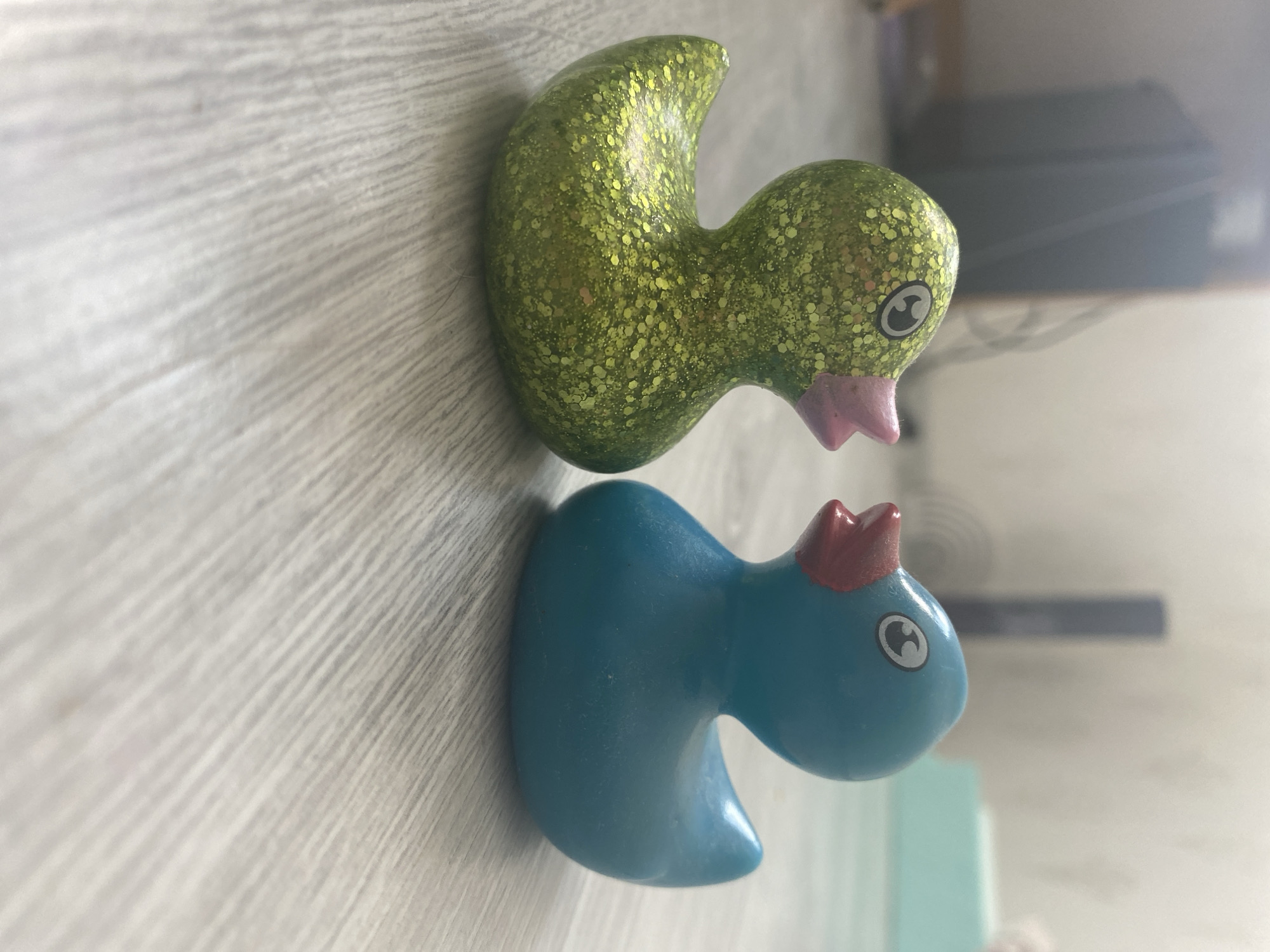 Two rubber ducks are positioned on a light-colored wooden floor, facing each other. The duck on the left is green and covered in glitter, while the duck on the right is solid blue. Both ducks have pink beaks and black and white eyes. The background is blurred, showing a light-colored wall and some indistinct objects. The scene appears to be indoors, likely in a domestic setting. There is no indication of the location being in Blagoevgrad, Bulgaria.