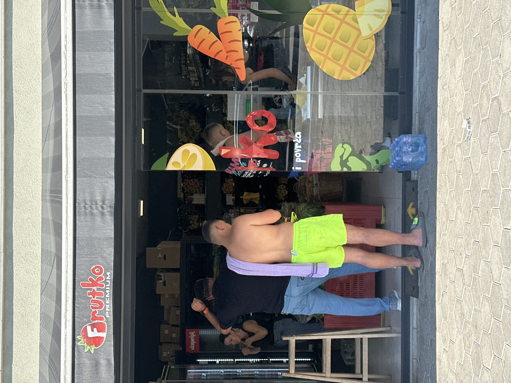 The image depicts the exterior of a shop named "Frutko PREMIUM" located in Budva, Montenegro. The shop's storefront features large glass windows adorned with colorful decals of fruits and vegetables, including a strawberry with the shop's logo, carrots, a slice of lemon, and a pineapple. Inside the shop, products are displayed on shelves, and a checkout counter is visible.
Two men and a woman are present. One man, shirtless and wearing neon green shorts, stands facing away from the camera, with a purple towel draped over his shoulder. Another man, wearing a black t-shirt and blue jeans, also faces away, with his arm raised. A woman in a black tank top and jeans stands near the entrance, to the left, with her hands in a boxing-like stance.
Outside the shop, on the paved sidewalk, several cases of bottled water are stacked. The ground is paved with hexagonal tiles. The overall scene suggests a sunny day, with the shop appearing to be open for business.