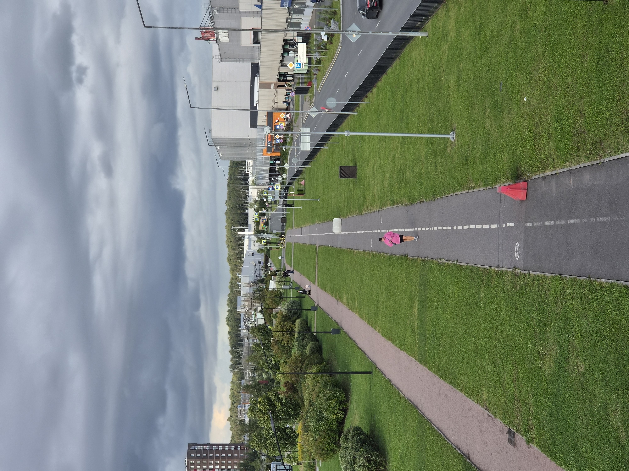 An overhead shot shows a person wearing a bright pink jacket and black shorts running on a paved path that is bordered by a grassy area. To the left of the path is a dirt path. In the background, there are trees, buildings, and a cloudy sky. The scene appears to be in a park-like area in Kudrovo, Russia, with commercial buildings and a residential building in the distance. There is a black car on the road to the right of the grassy area. A red traffic cone is visible near the running path.