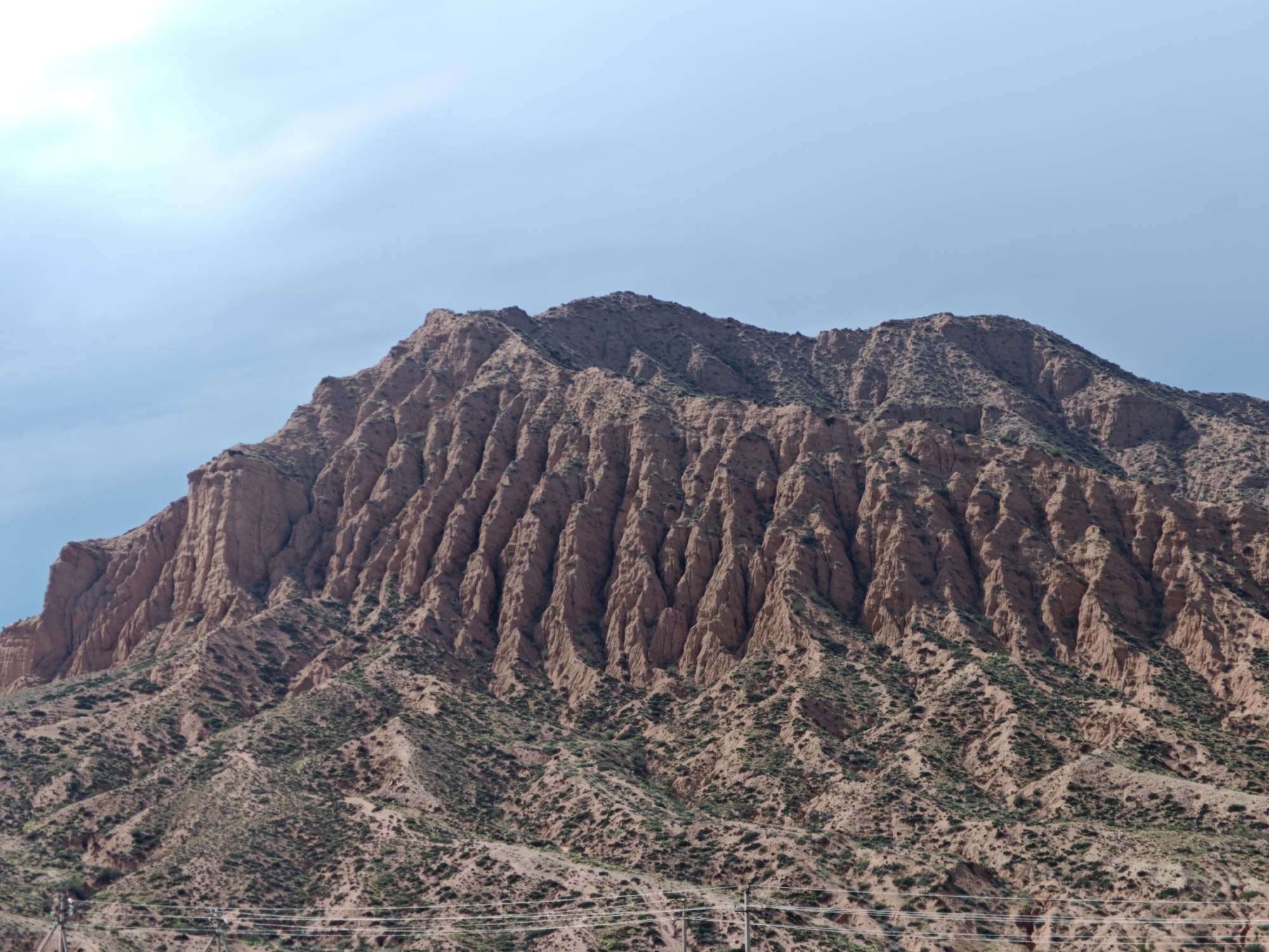 The image shows a close-up, low-angle shot of a large, eroded, reddish-brown mountain in Kadji Say, Kyrgyzstan. The mountain has steep, deeply grooved sides, with visible stratification and a rough, uneven texture. Sparse green vegetation, appearing as low shrubs or grasses, is scattered across the lower slopes and in some of the crevices on the mountain's face. The sky above is a clear, pale blue with some faint wisps of clouds on the left side. In the lower portion of the image, a few power lines and utility poles are visible, suggesting the presence of infrastructure near the mountain's base. The overall scene is natural and geological, with a focus on the imposing natural formation.