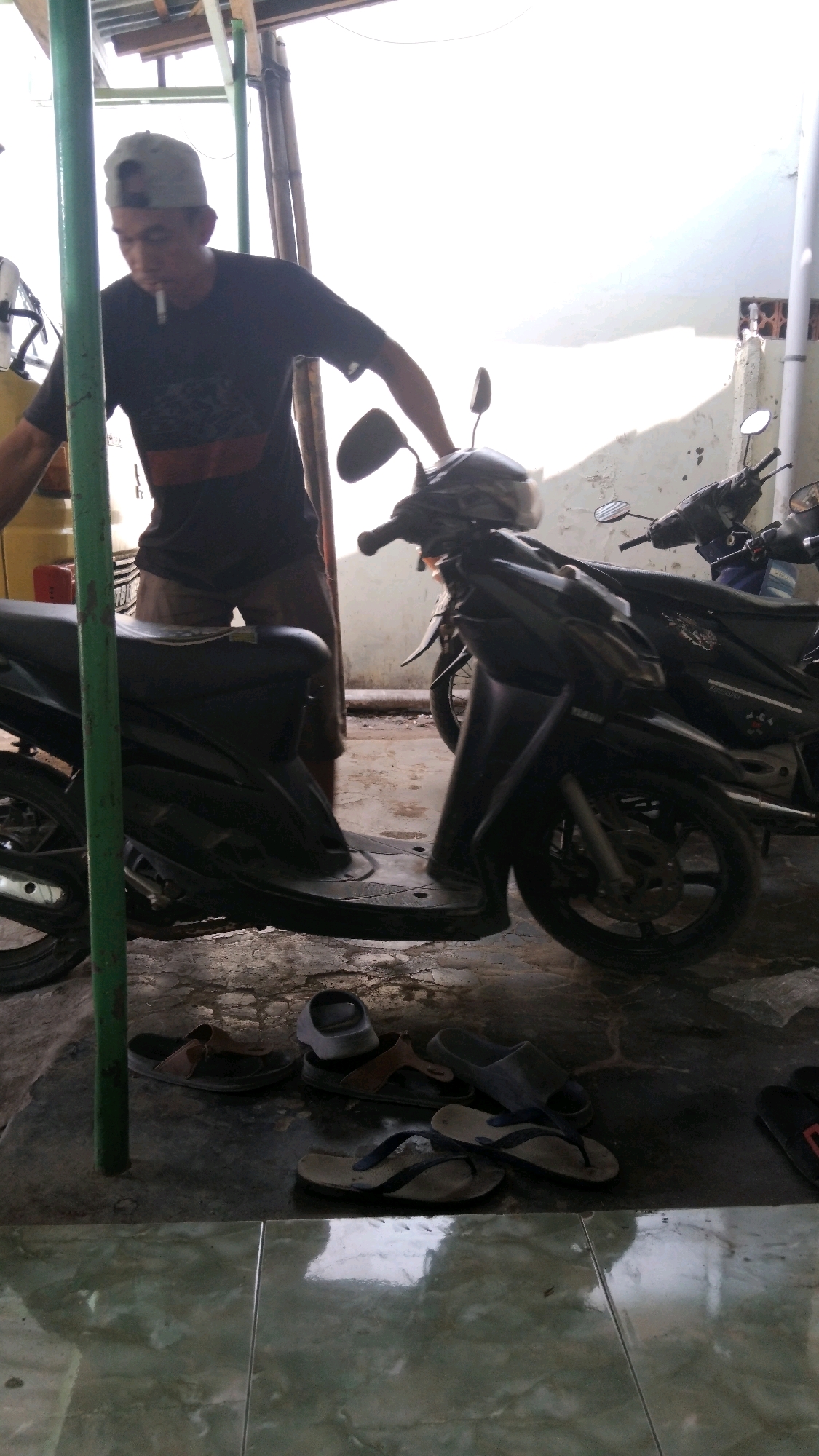 A man wearing a dark t-shirt and a grey baseball cap is standing next to two motorcycles. He is smoking a cigarette and appears to be working on or near the motorcycles. The motorcycles are parked on a concrete floor, with several pairs of sandals scattered around them. The scene is outdoors, with a bright, overcast sky suggesting it might be daytime. The environment appears to be a workshop or a parking area, possibly in a residential or commercial area of Dukuhturi, Indonesia. The overall impression is one of a casual, everyday scene.