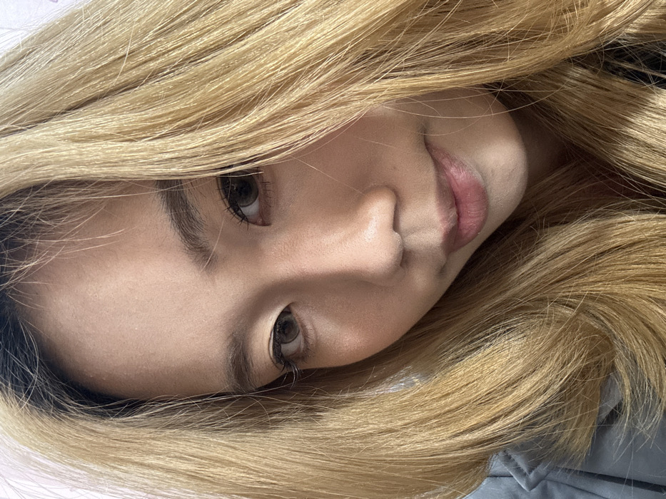 A close-up portrait of an East Asian female, taken in Shapingba District, China, occupies the majority of the frame. The subject's face is presented directly towards the viewer, slightly angled. She possesses long, wavy hair, dyed blonde with distinct dark roots visible at the scalp. Her eyes are dark brown, exhibiting a clear iris pattern, and her eyebrows are defined. The subject's nose is centrally located on the face, and her lips are softly closed in a neutral expression. A light-colored garment, appearing grey or light blue, is visible on her left shoulder and upper arm. The background is out of focus and lacks specific discernible features.