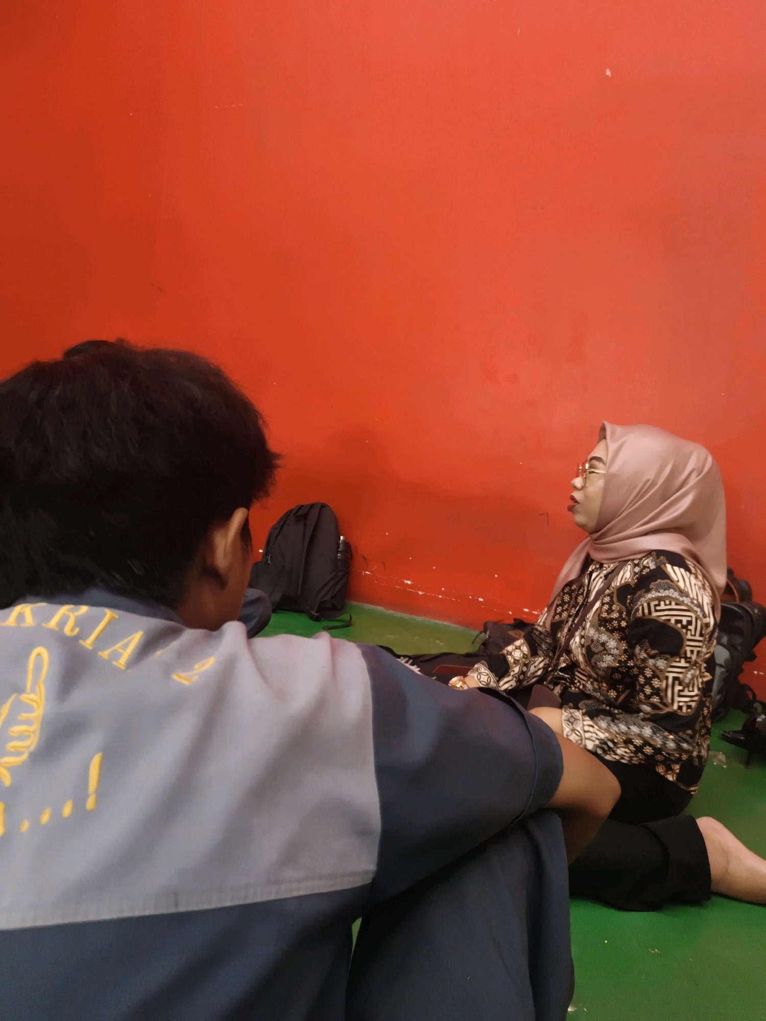 In Katrungan, Indonesia, two individuals are seated on a green floor in front of a vibrant red wall. The person in the foreground, viewed from behind, has dark hair and is wearing a grey and dark blue jacket with yellow lettering. To their right, a woman wearing a light pink hijab, glasses, and a patterned black and white top is also seated. A black backpack is visible behind her. The scene suggests a moment of rest or conversation in an indoor setting.