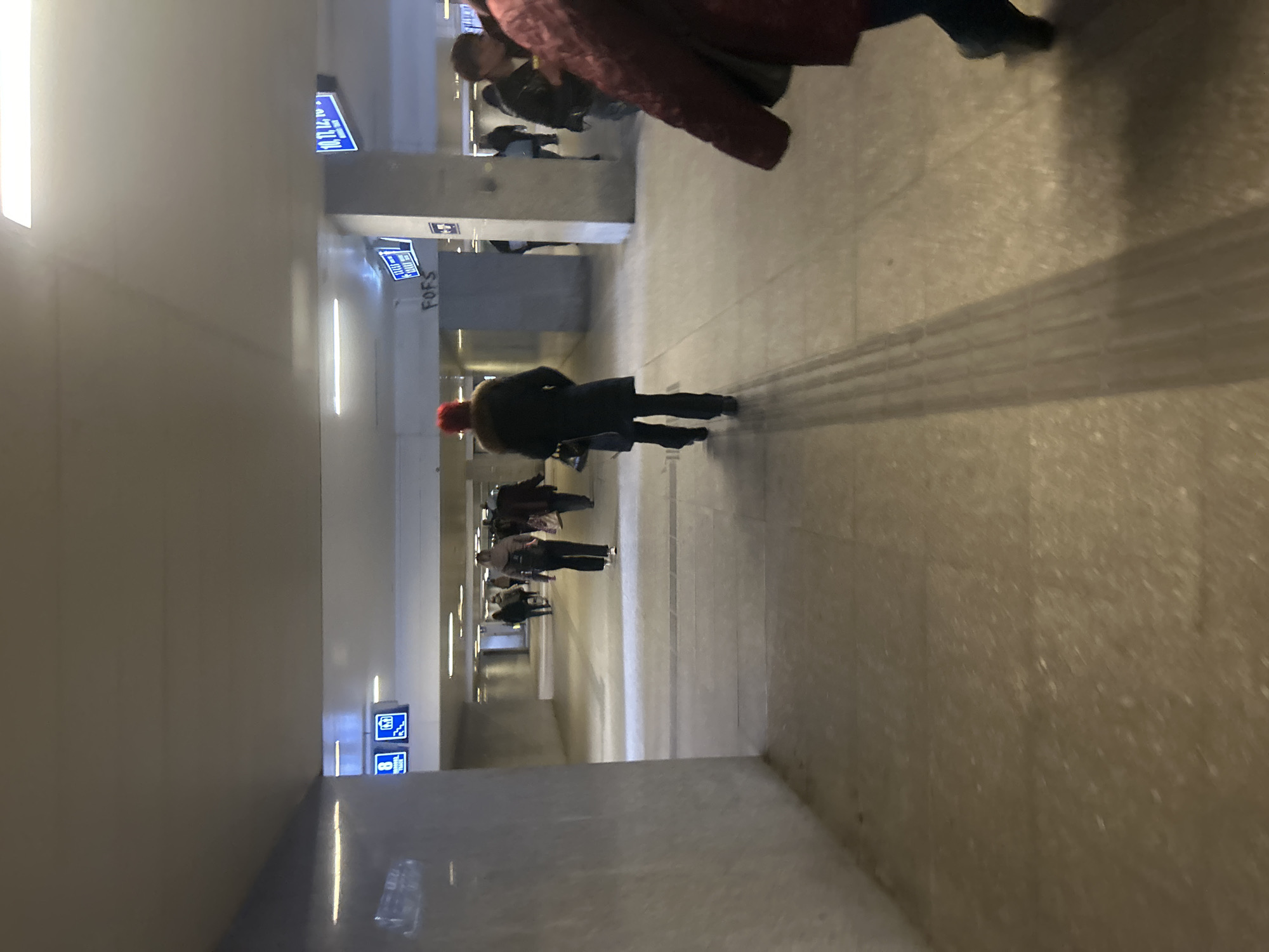 The image depicts an indoor pedestrian walkway in Sofia, Bulgaria, characterized by a minimalist architectural style with concrete walls and ceiling, interspersed with recessed lighting fixtures. A series of blue and white informational signs are affixed to the walls.
Several individuals are present, walking in both directions. A person wearing a dark coat and a red hat is moving away from the viewer, while others are walking towards the viewer. The perspective suggests a point of view slightly above ground level, potentially from a standing position. The flooring appears to be made of concrete or a similar material.