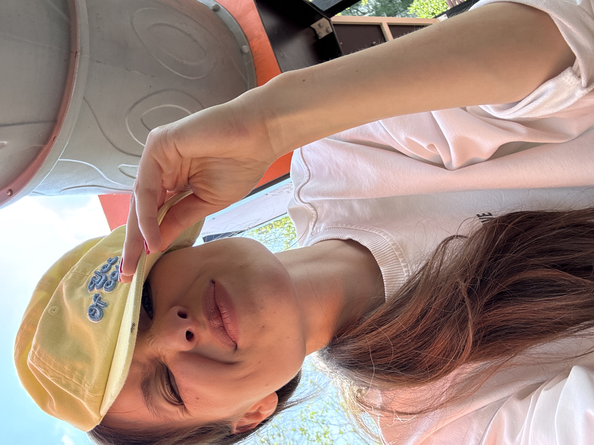 A young woman wearing a yellow cap and a white t-shirt is positioned in front of a gray and orange metallic structure, possibly a playground feature. Her head is tilted back, and she is holding the brim of her cap with her hand. The background shows glimpses of green foliage, suggesting an outdoor setting. The location is likely a park in the city of Moscow, Russia.