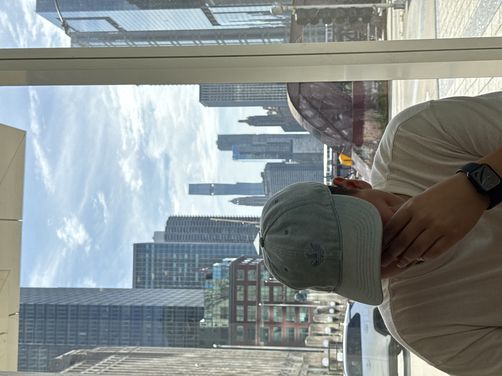 A person wearing a light blue baseball cap and a cream-colored t-shirt is seated indoors, with their hand covering their face. They are looking out a window, which offers a view of the Chicago cityscape. The window frame is visible on the right side of the image, and the city skyline features numerous tall buildings, a cloudy sky, and a visible arched structure, likely a bridge or elevated roadway. A white SUV is parked outside the window to the left. The scene appears to be a daytime setting in an urban environment.