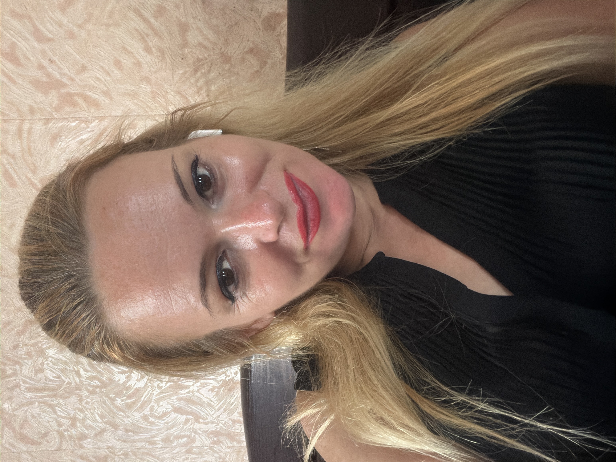 A woman with blonde hair is pictured in a close-up shot, wearing a black top. She is smiling, and her lips are painted red. She has on white earbuds. The background is a textured, light-colored wall. The image appears to be a selfie taken indoors.