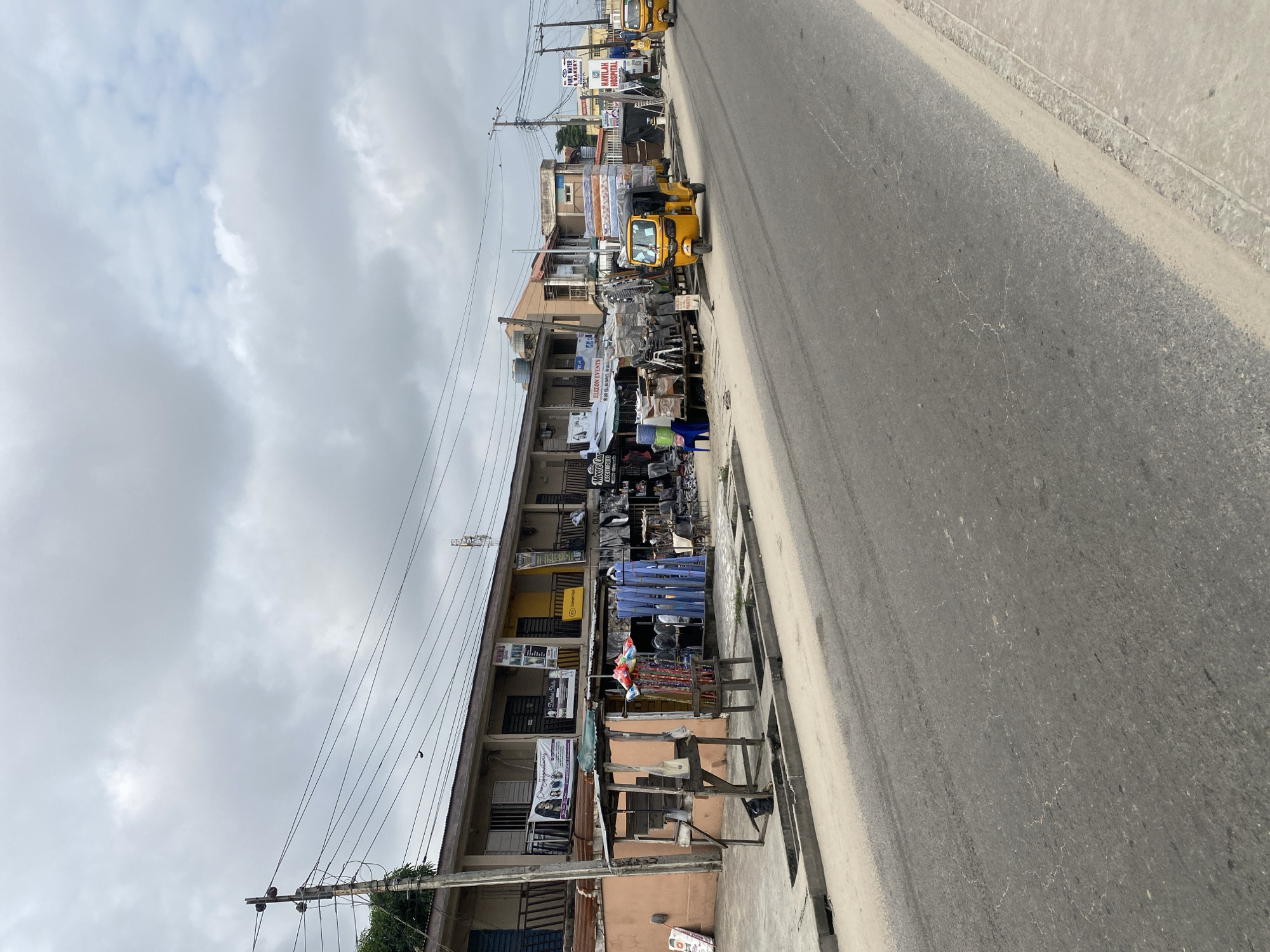 The image captures a street scene in a city with a cloudy sky overhead. The street is paved with asphalt, showing some signs of wear and tire marks. A concrete drainage system runs along the side of the road. On the left side of the street, a row of shops and businesses are visible. These buildings have multiple stories with balconies and exposed electrical wiring running overhead. In front of the shops, there are various stalls and displays of goods. One stall features wooden structures with items like brooms and what appear to be household goods for sale. The shops themselves display banners and signs, some of which are partially obscured. One sign reads "Moore Car Accessories." Another advertises "ELIZON EVENTS." Further down the street, a yellow auto-rickshaw, also known as a tuk-tuk, is parked by the roadside. Behind it, another similar vehicle is seen moving away. Another sign indicates "PURE WATER BAKERY" and "HAVILAH HOSPITAL." The overall impression is of a bustling commercial area in a developing urban environment.