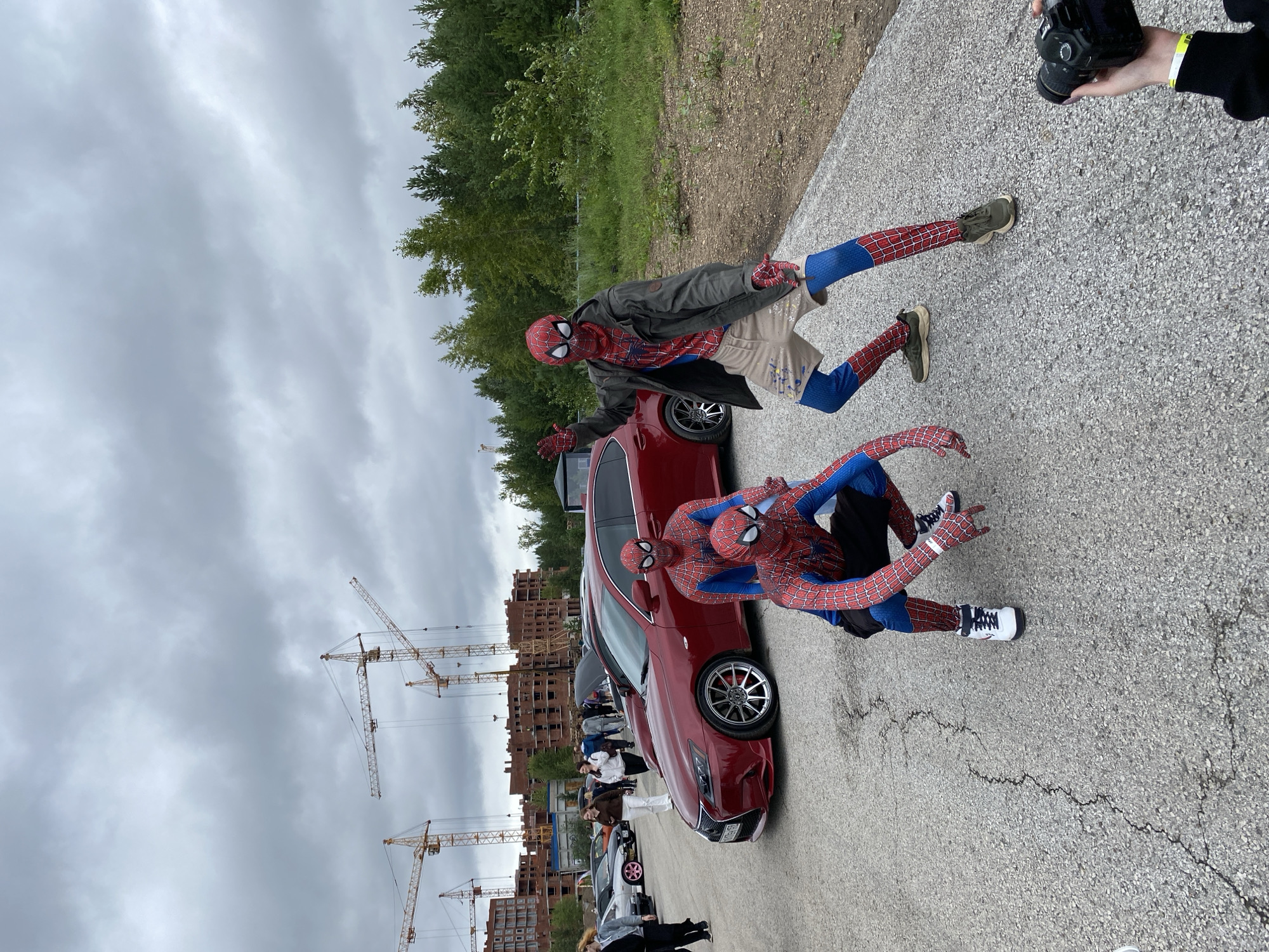 Three individuals in Spider-Man costumes are posing on a paved area in front of a red Lexus sedan. The person on the right is standing with one hand raised, wearing a jacket and shorts over the costume. The other two are in a crouching position. In the background, several construction cranes and unfinished brick buildings are visible, indicating a construction site. A person's hand holding a camera is in the lower right corner of the frame. The sky is overcast. The location is identified as Perm, Russia.